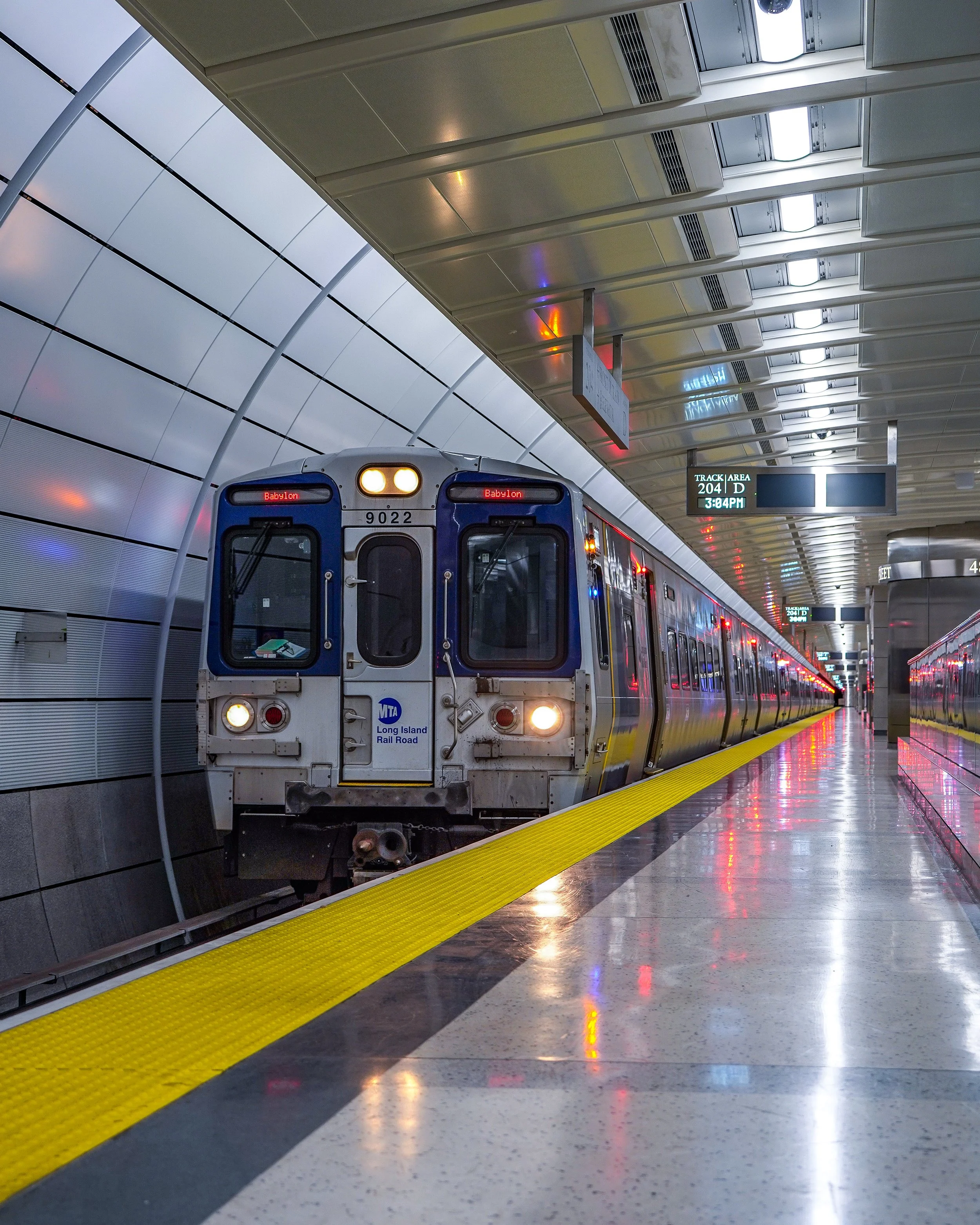 An MTA Long Island Rail Road train at an underground subway station, with the sign indicating it's heading to Babylon, passing through track D at 3:04 PM.