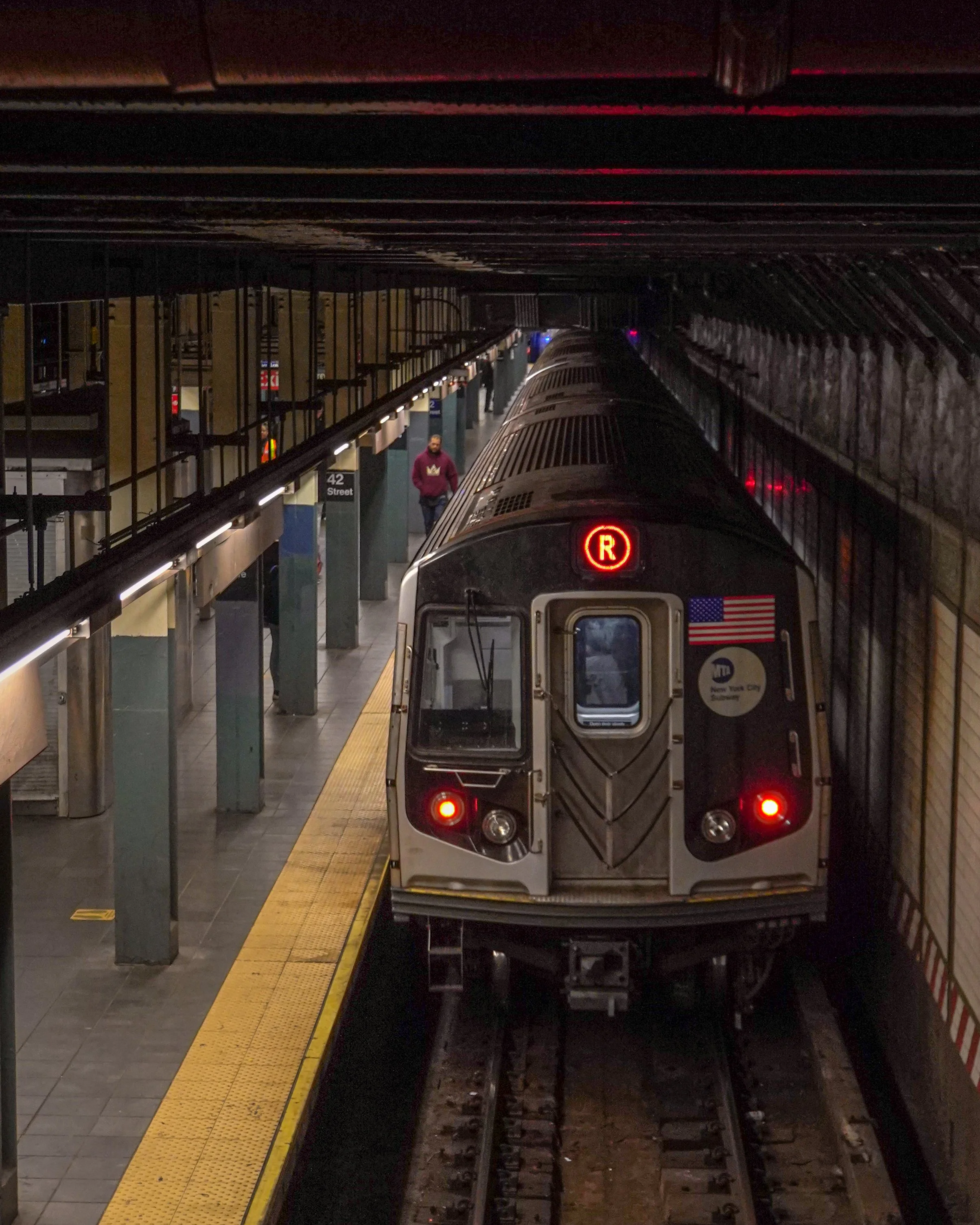 An NYC subway train departing Times Square platform in a subway station with a few passengers waiting.