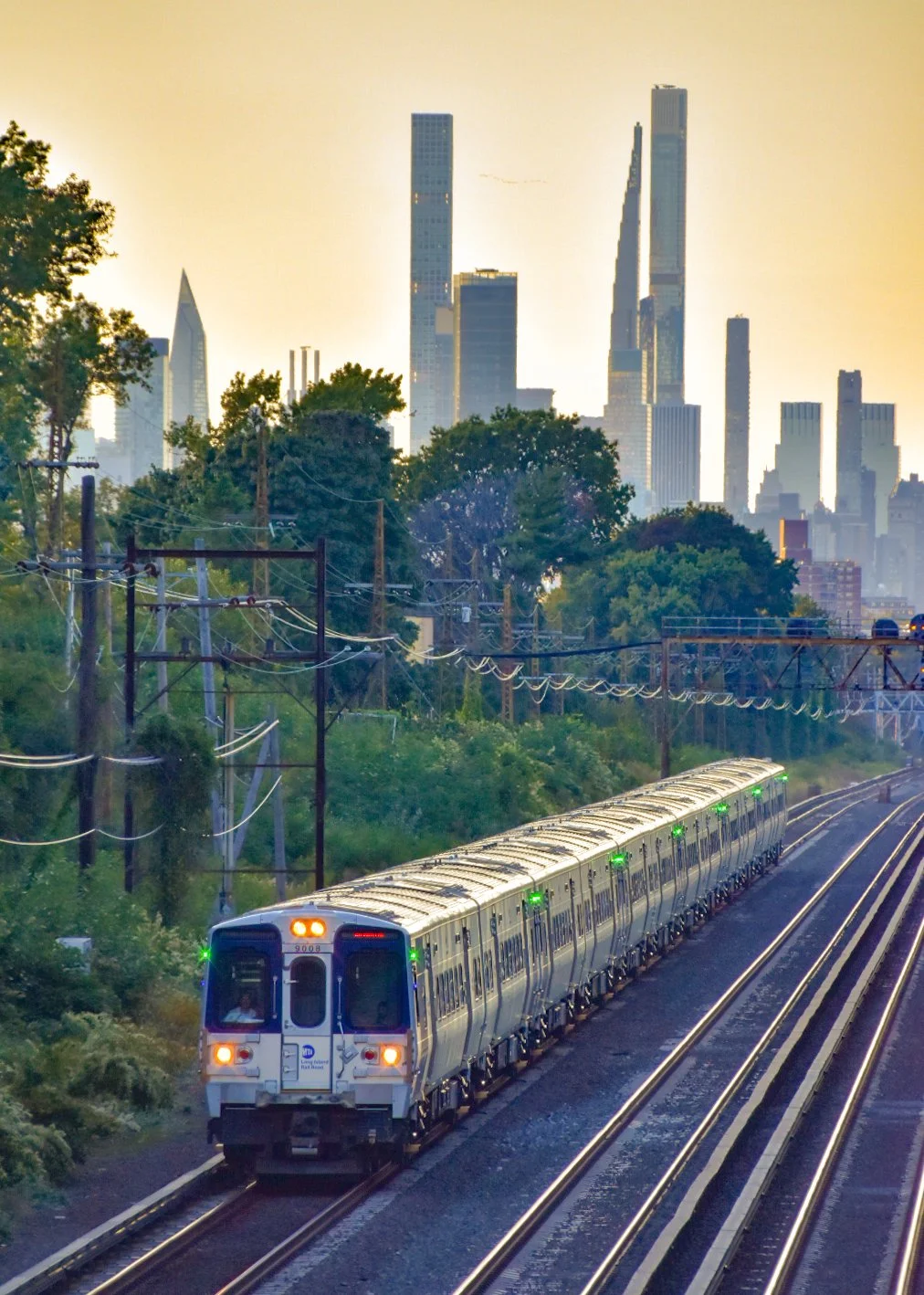 A city train traveling on elevated tracks with a skyline of tall modern skyscrapers in the background, green trees, and utility poles along the tracks at sunset or sunrise.