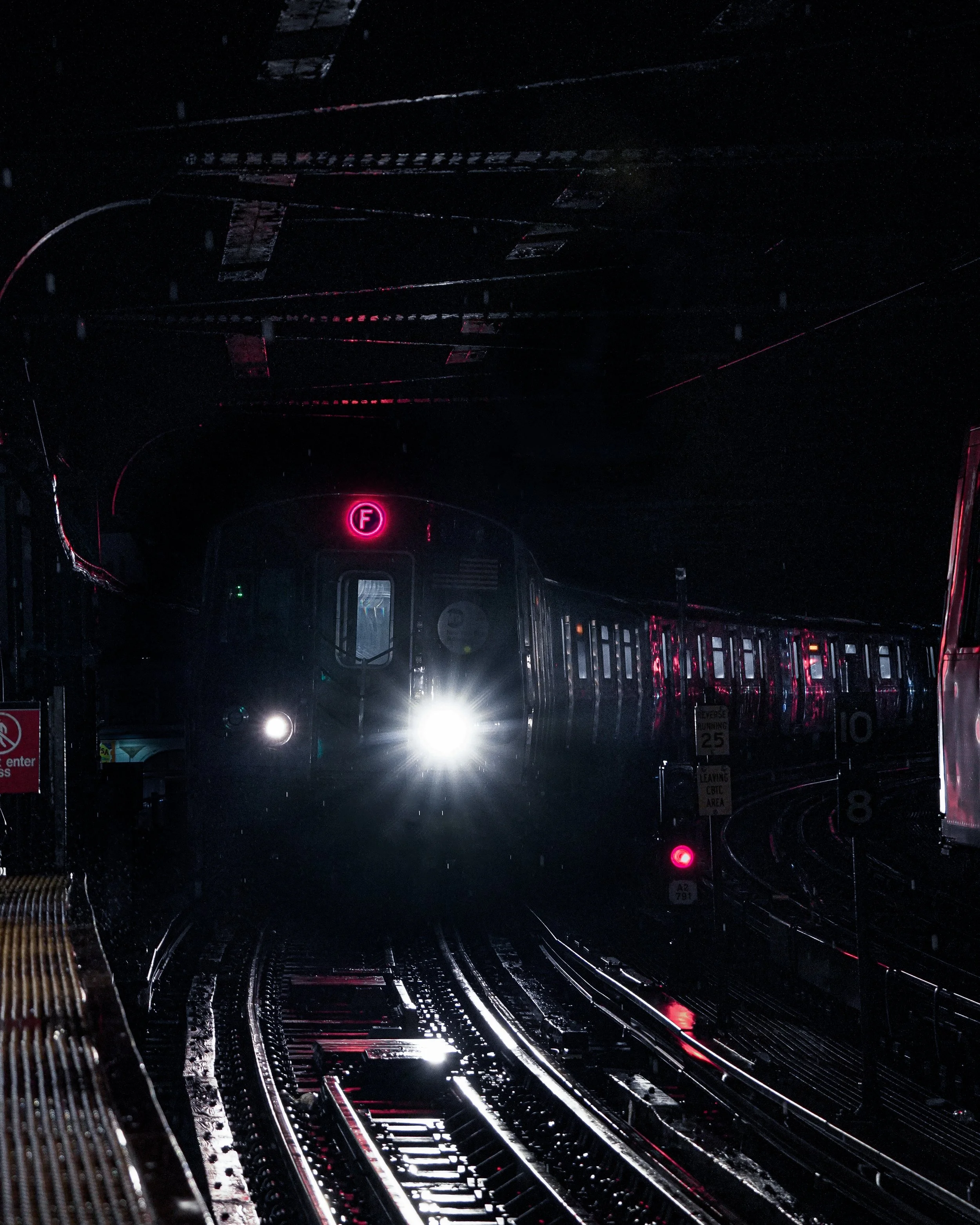 A subway train emerging from the tunnel in a dark underground station, with its headlights on and a pink 'F' sign illuminated on the front.