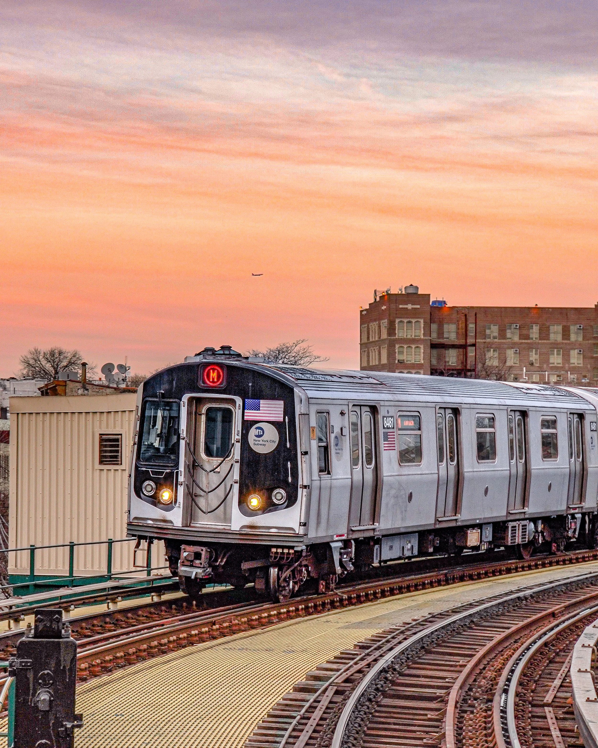 A New York City subway train on an elevated track during sunset with a colorful sky in the background.