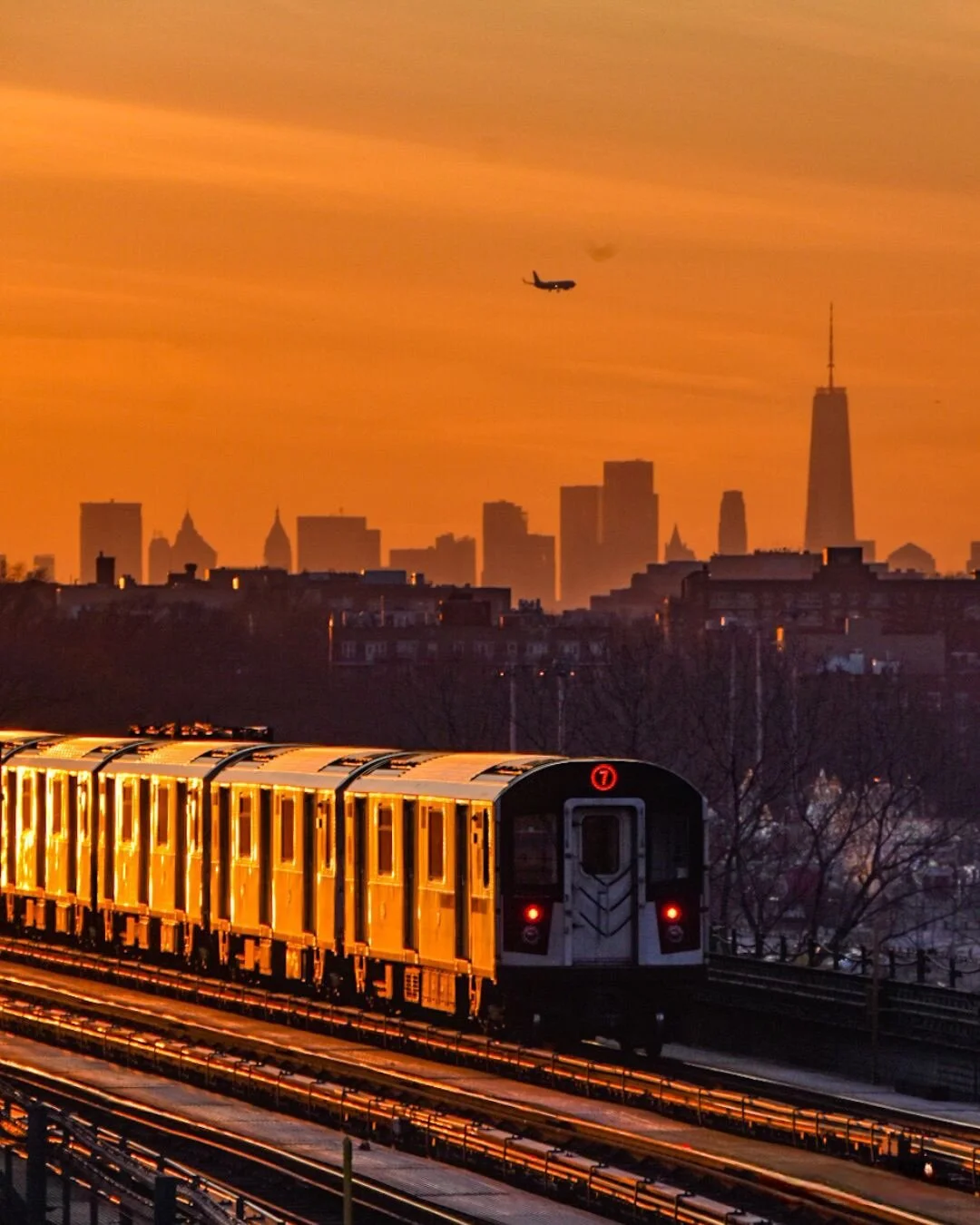 Sunset over New York City skyline with an airplane flying above and a yellow subway train on an elevated track in the foreground.