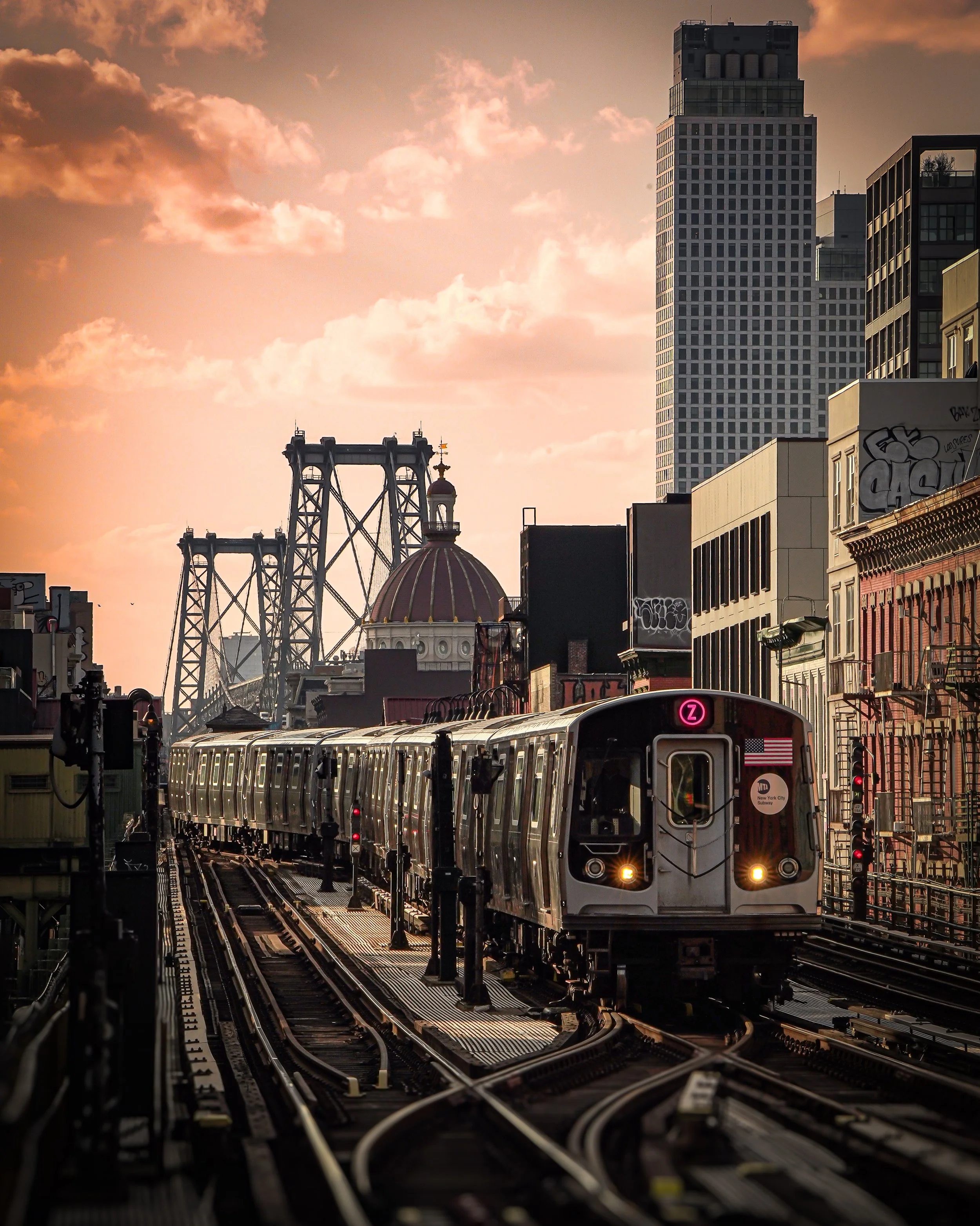 An elevated city train moving along tracks with city buildings and the Williamsburg Bridge in the background during sunset.