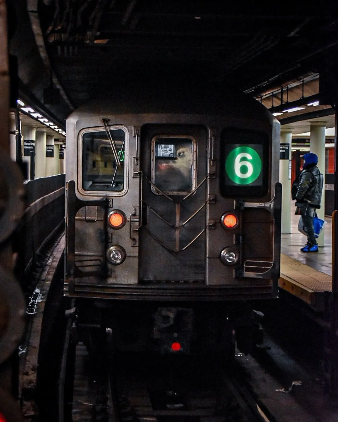 Front view of a subway train with a green number 6 display on a dimly lit underground station platform, with a person to the right standing and holding a shopping bag.