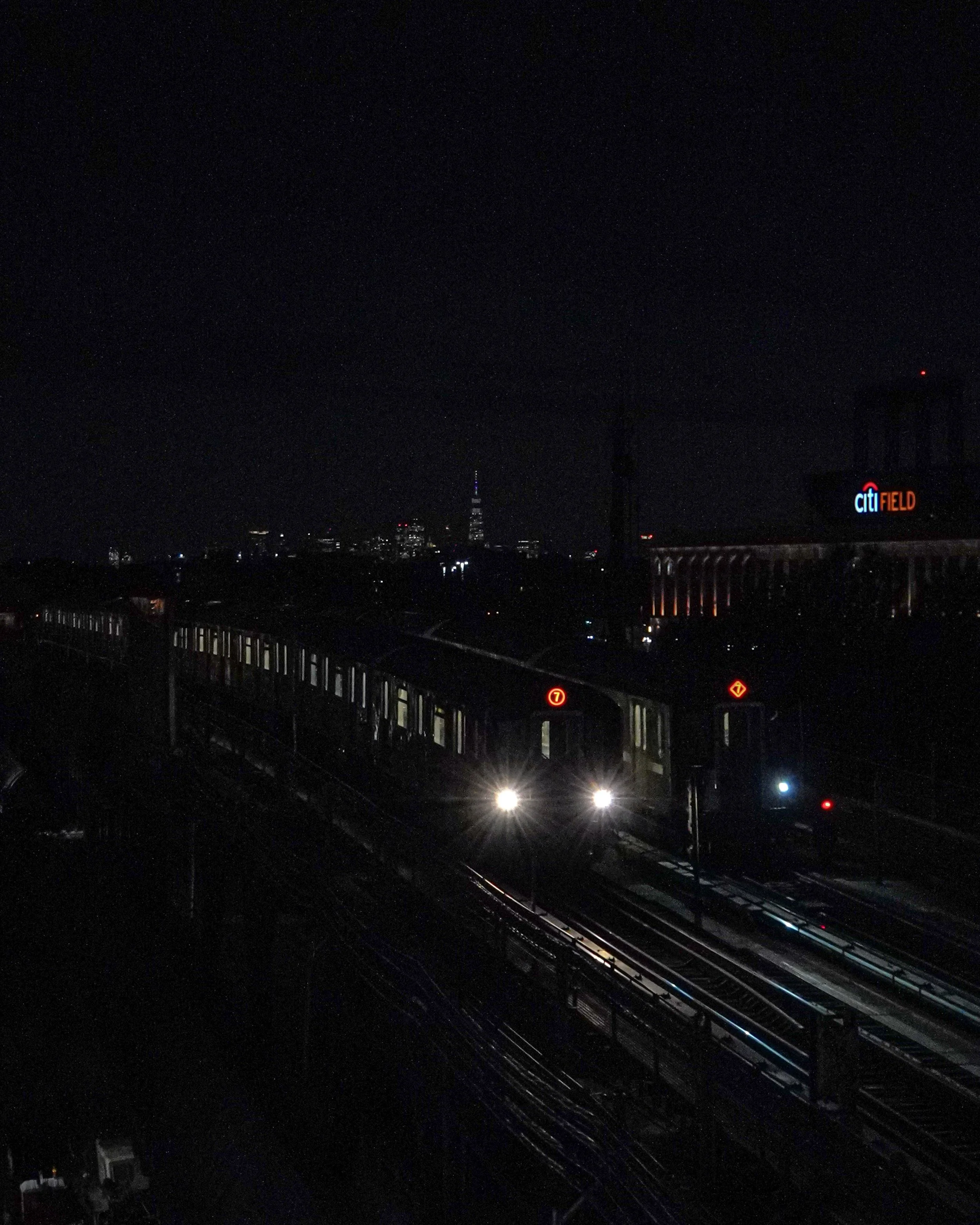Nighttime scene of a train on elevated tracks with city skyline in the background, including the Freedom Tower and a lit Citi Field sign.