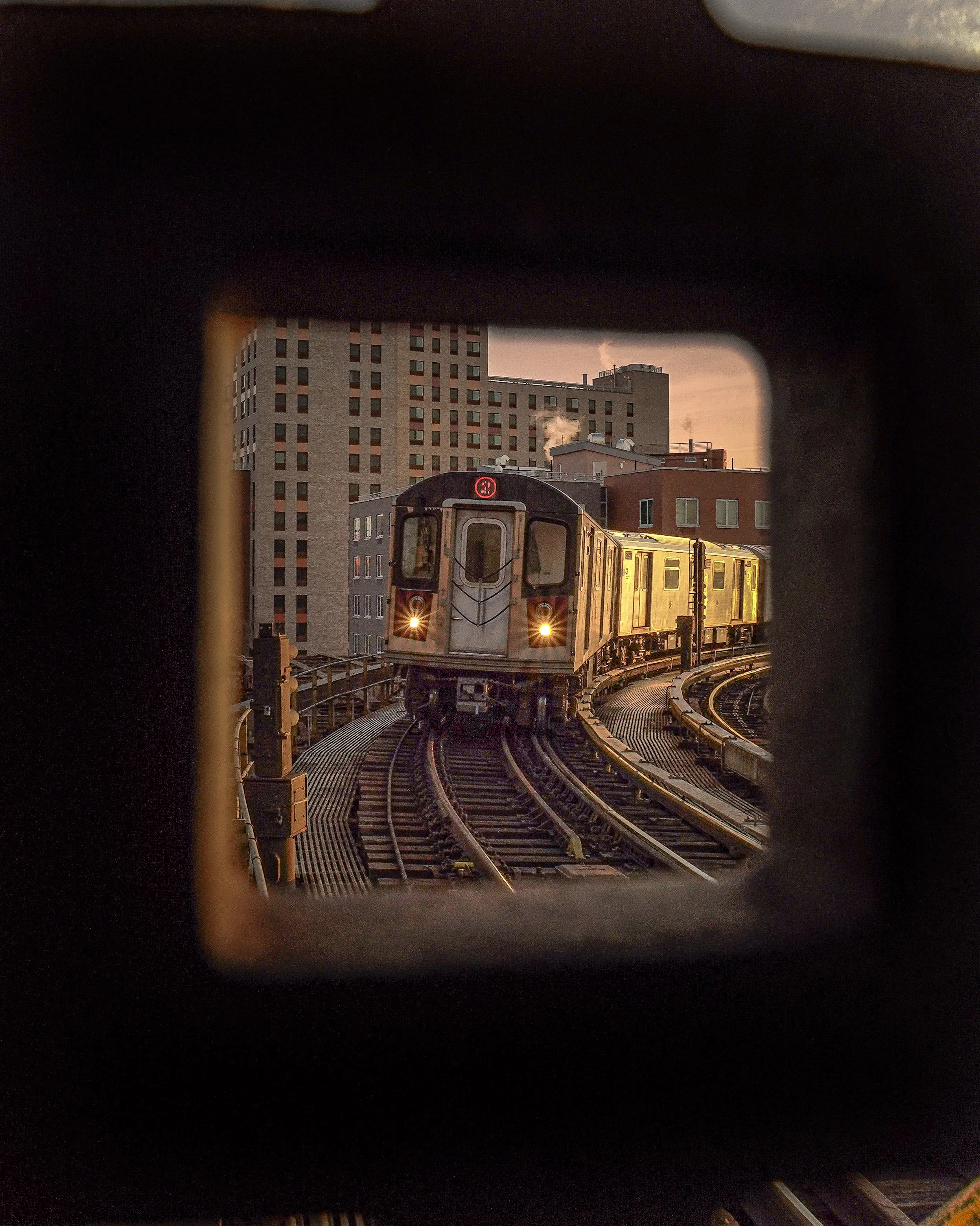 A train approaching on an elevated track during sunset, viewed through a square opening in a dark structure, with buildings in the background.