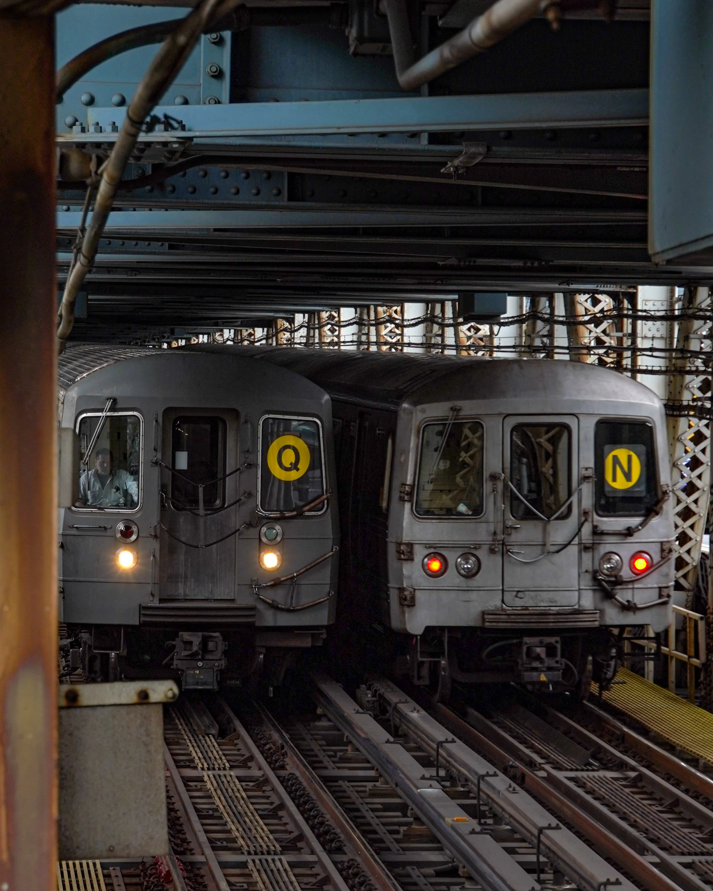 Two subway trains, one labeled 'Q' and the other 'N', are lined up on parallel tracks under a bridge or platform in an underground station.