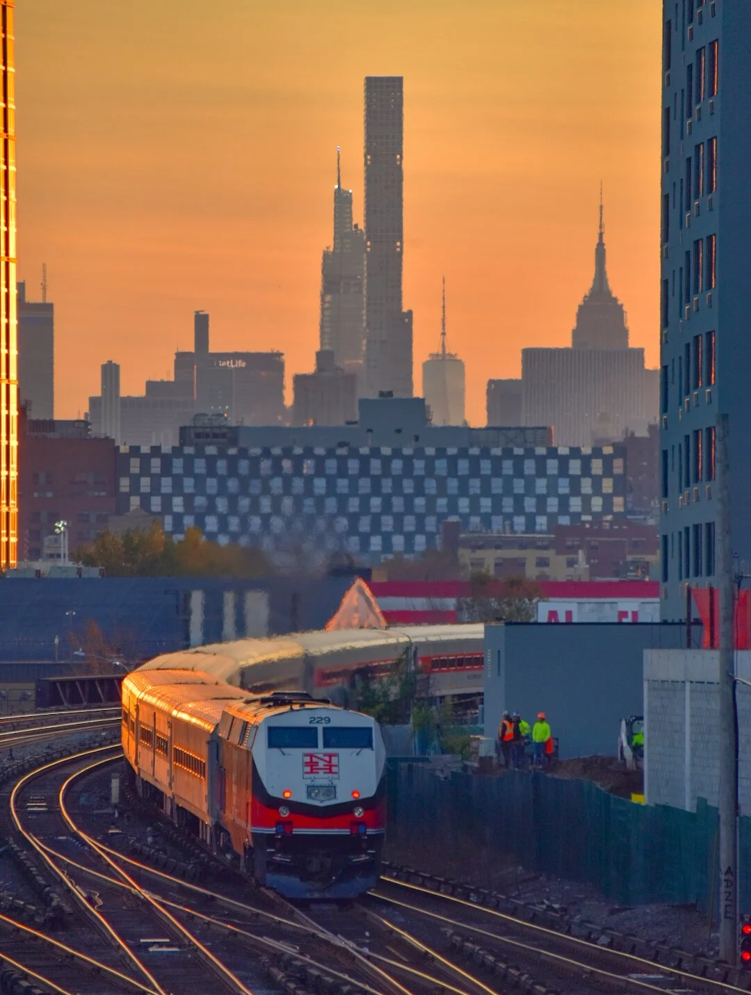 A train traveling on tracks in an urban cityscape at sunset, with high-rise buildings, the Empire State Building, and the city skyline in the background, and construction workers near the tracks.