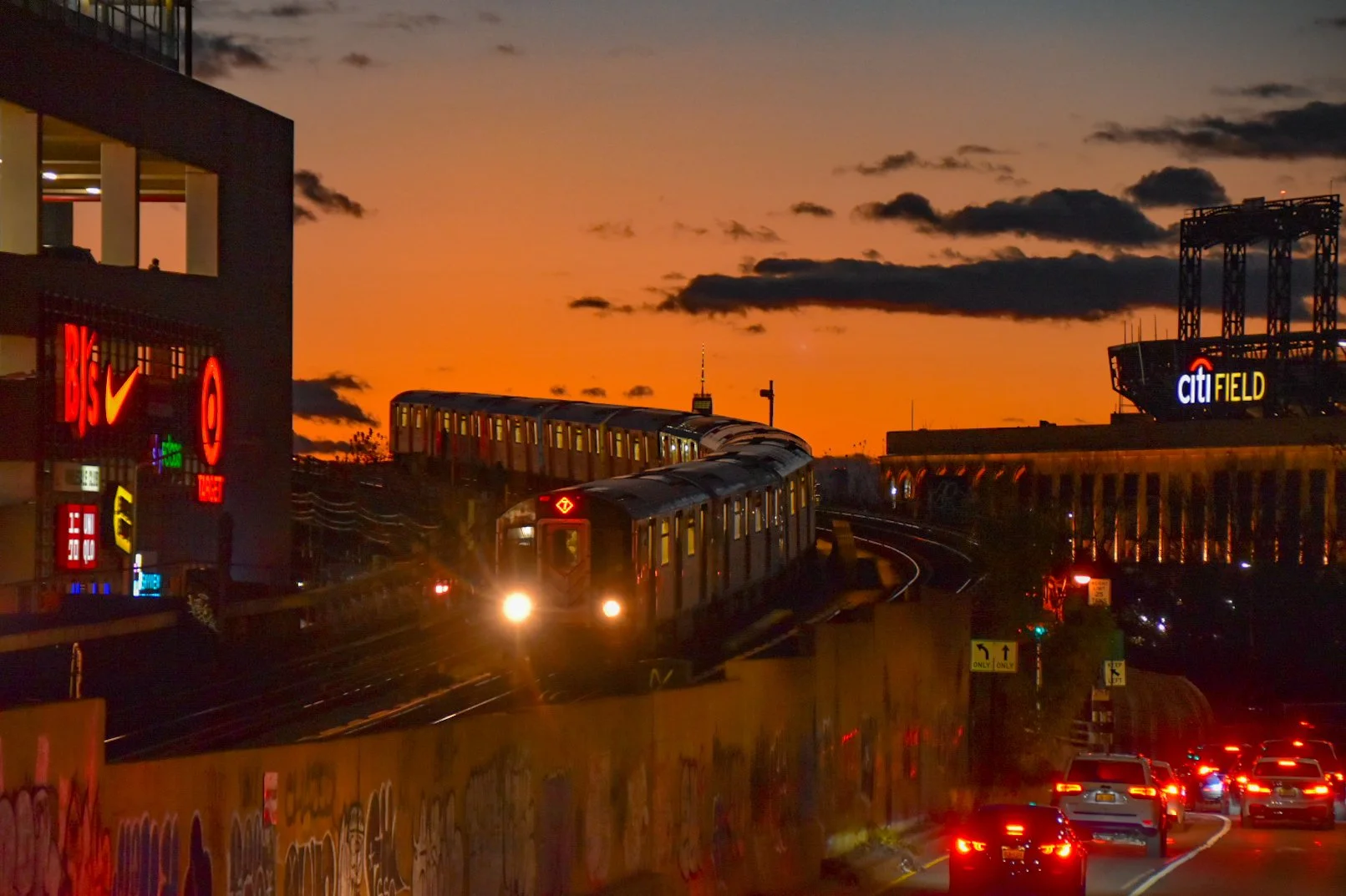 Subway train riding on an elevated track at sunset with city buildings, traffic, and signs in the background.