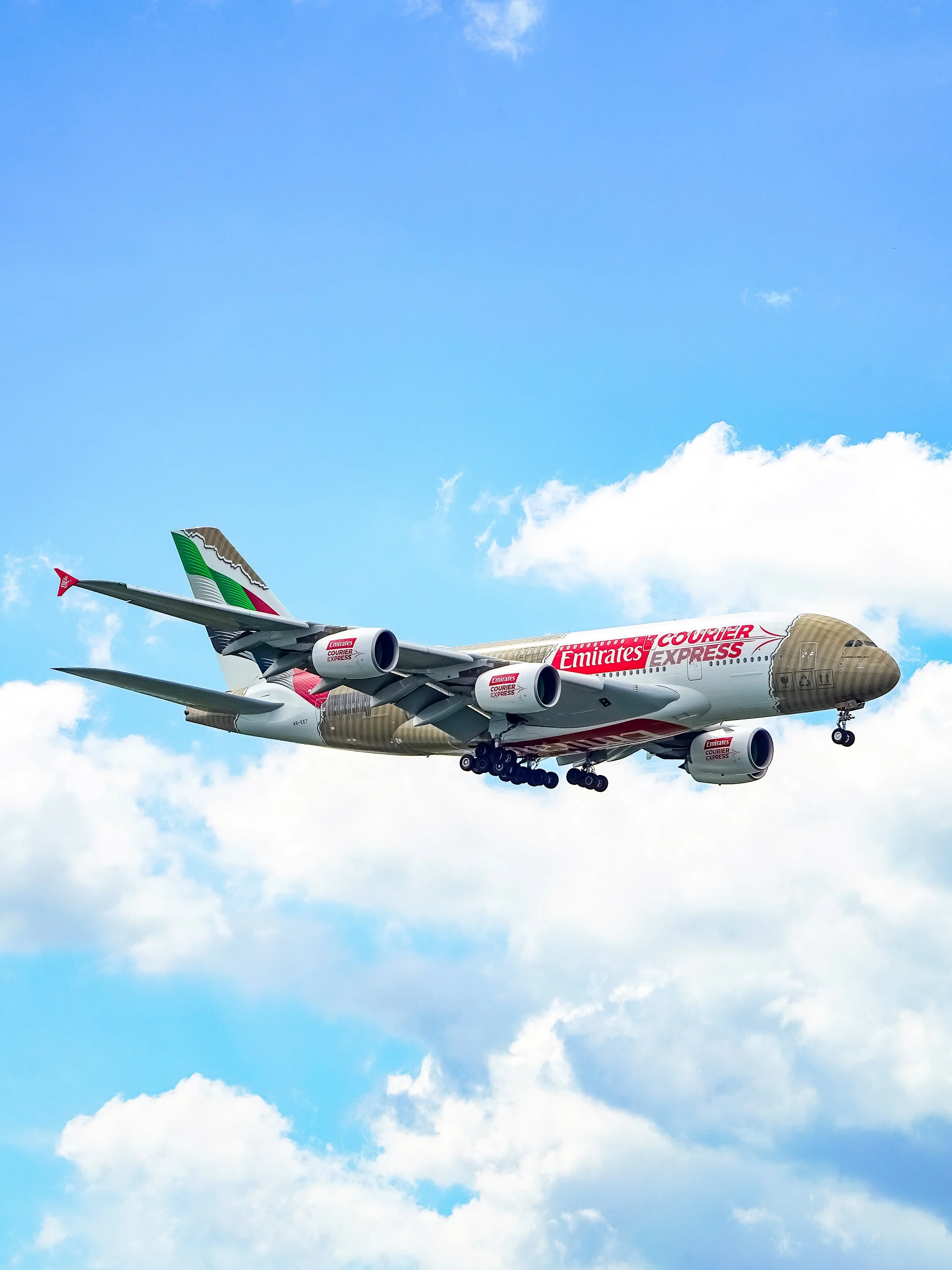 A large Emirates cargo airplane flying in a clear sky with some clouds, decorated with branding for Emirates Courier Express.