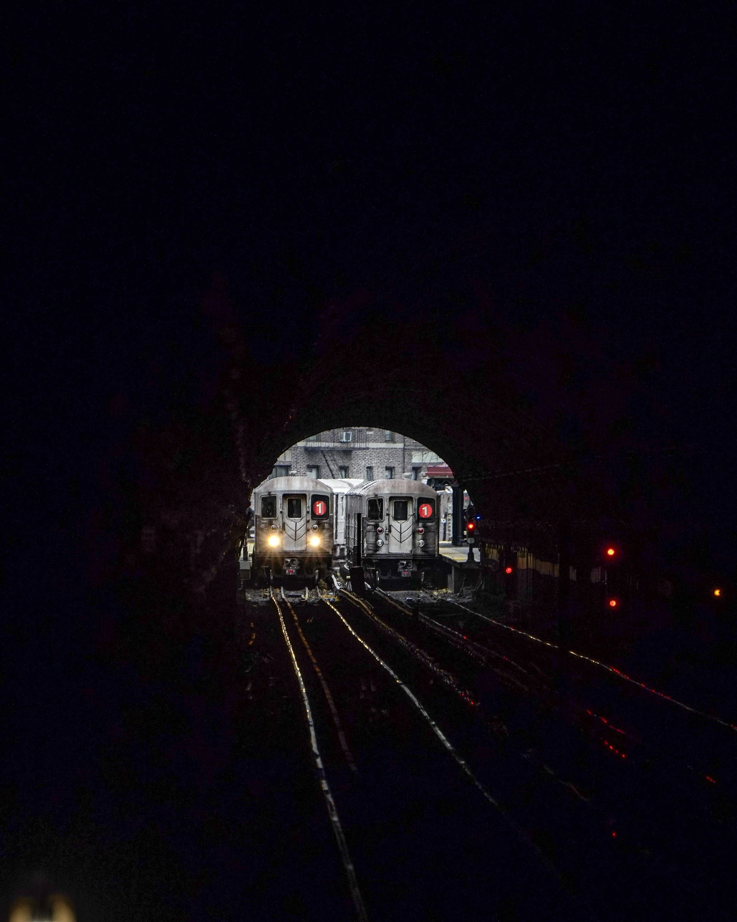 Two subway trains approaching a dark tunnel exit with city buildings in the background.