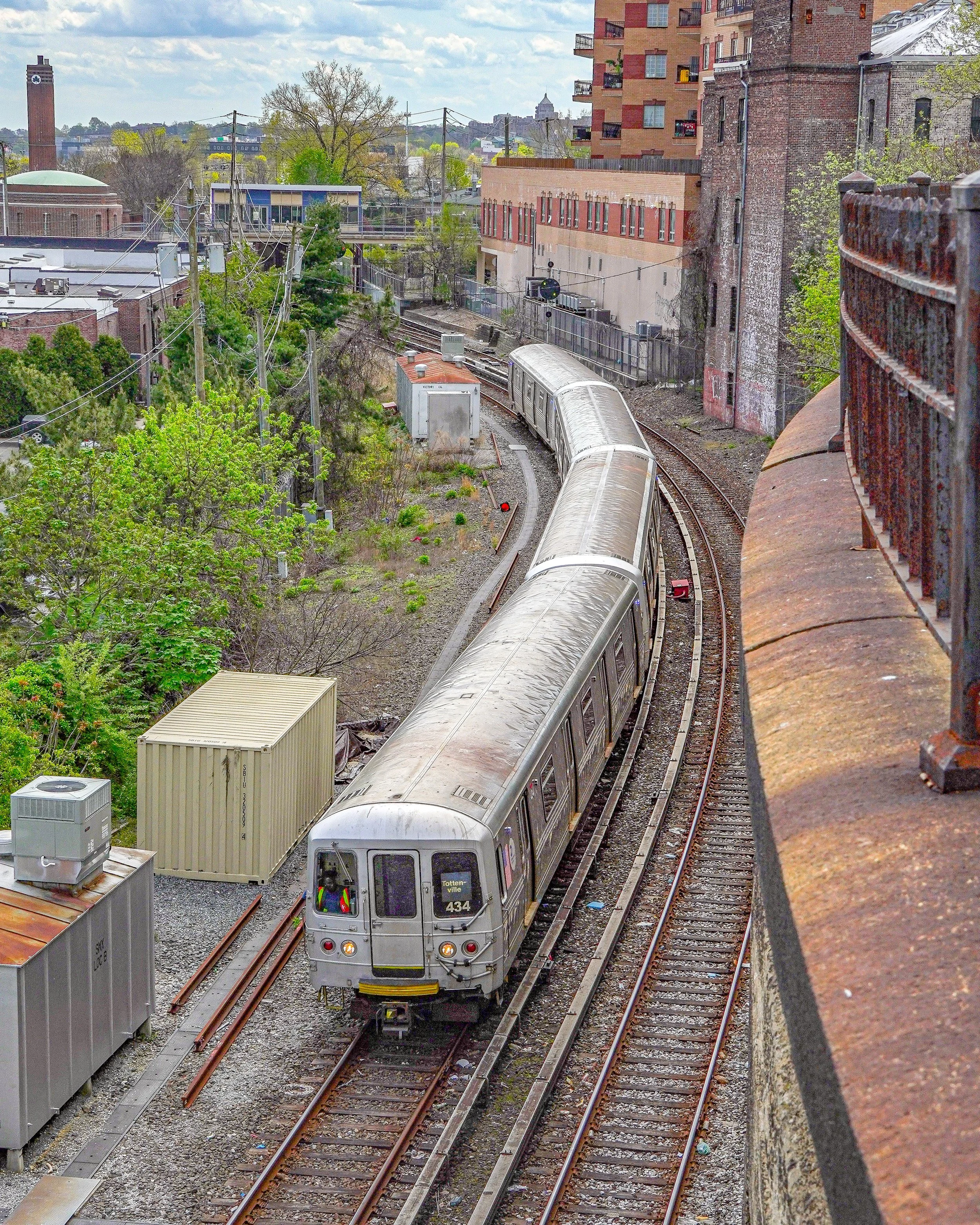 A silver light rail train traveling on curved tracks through an urban area with residential buildings, trees, and utility poles.