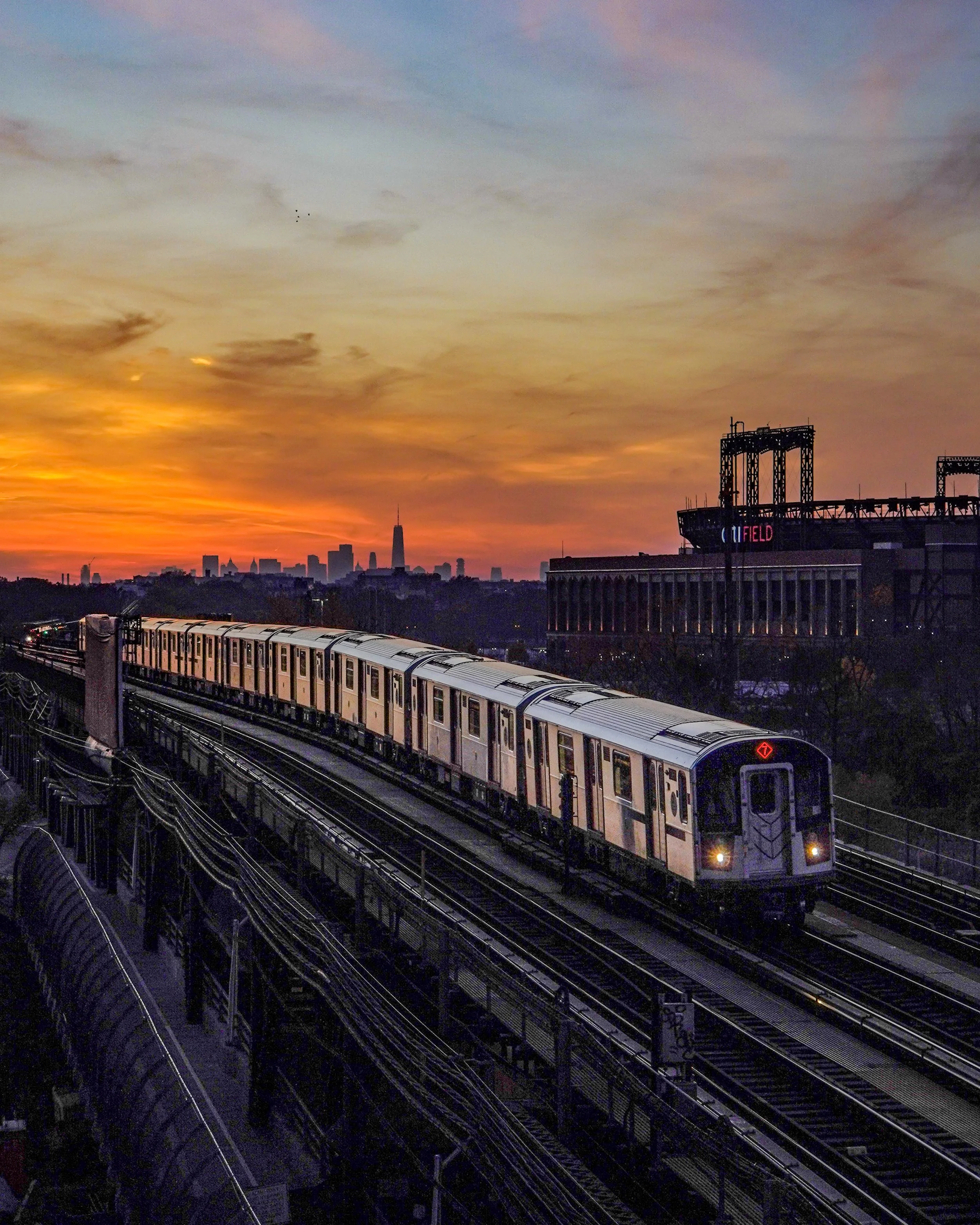 Subway train traveling on elevated tracks during sunset with city skyline in the background.