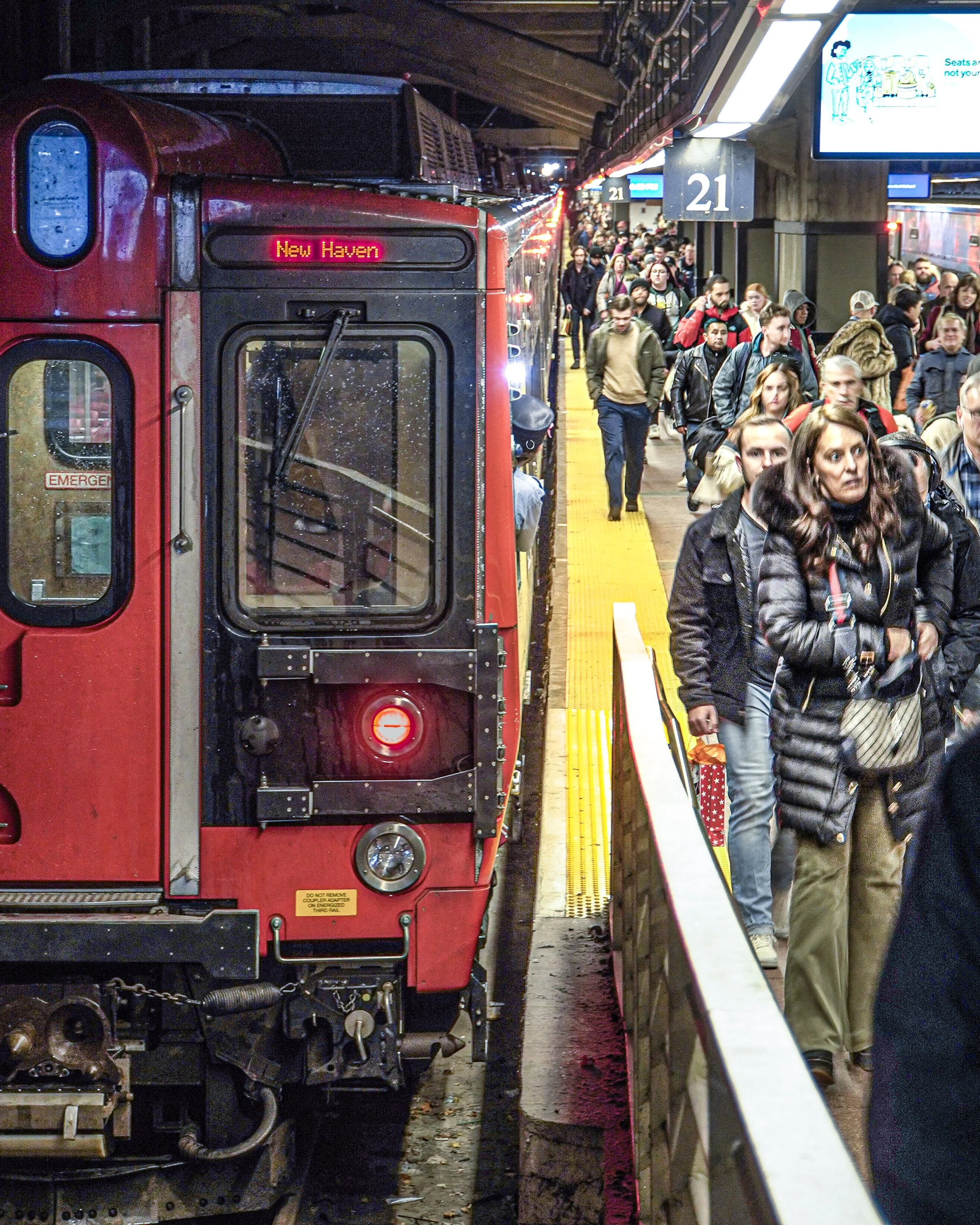 Crowded subway platform with a red train at the New Haven station, with people waiting to board and digital signs overhead.
