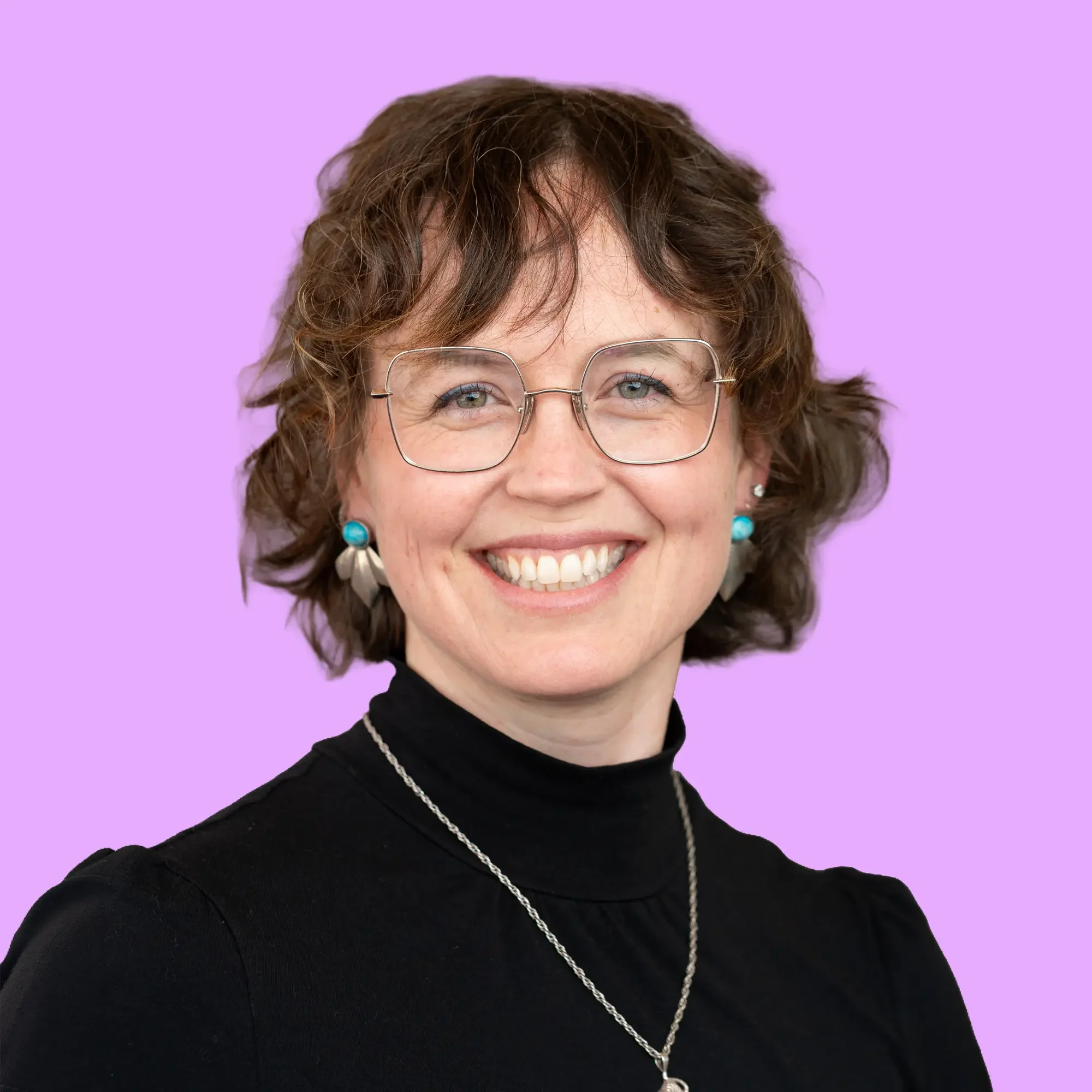 A woman faces the camera smiling for a headshot, she has short curly dark hair, glasses, a black turtleneck and a necklace