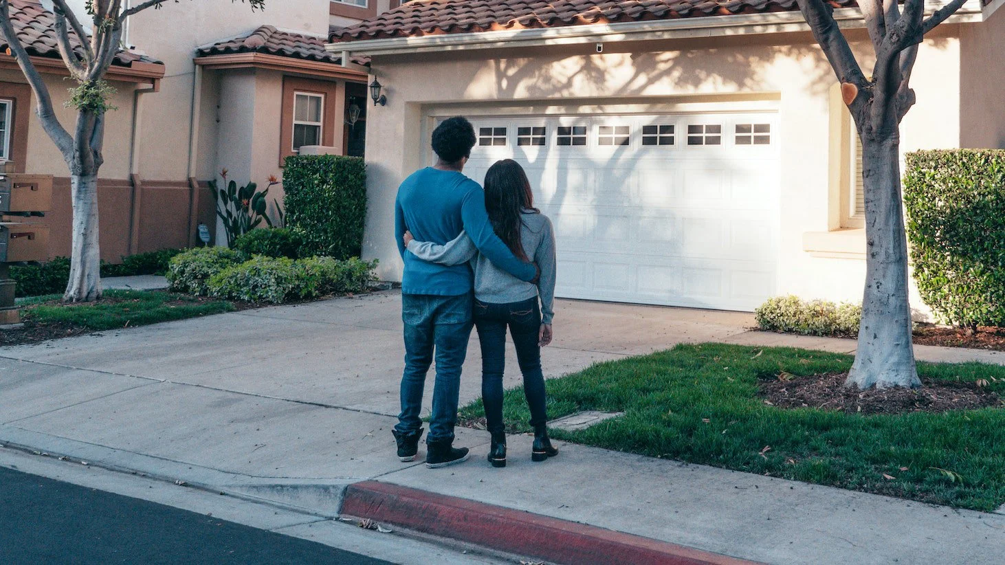 Two people stand in front of a home together with their arms supporting one another
