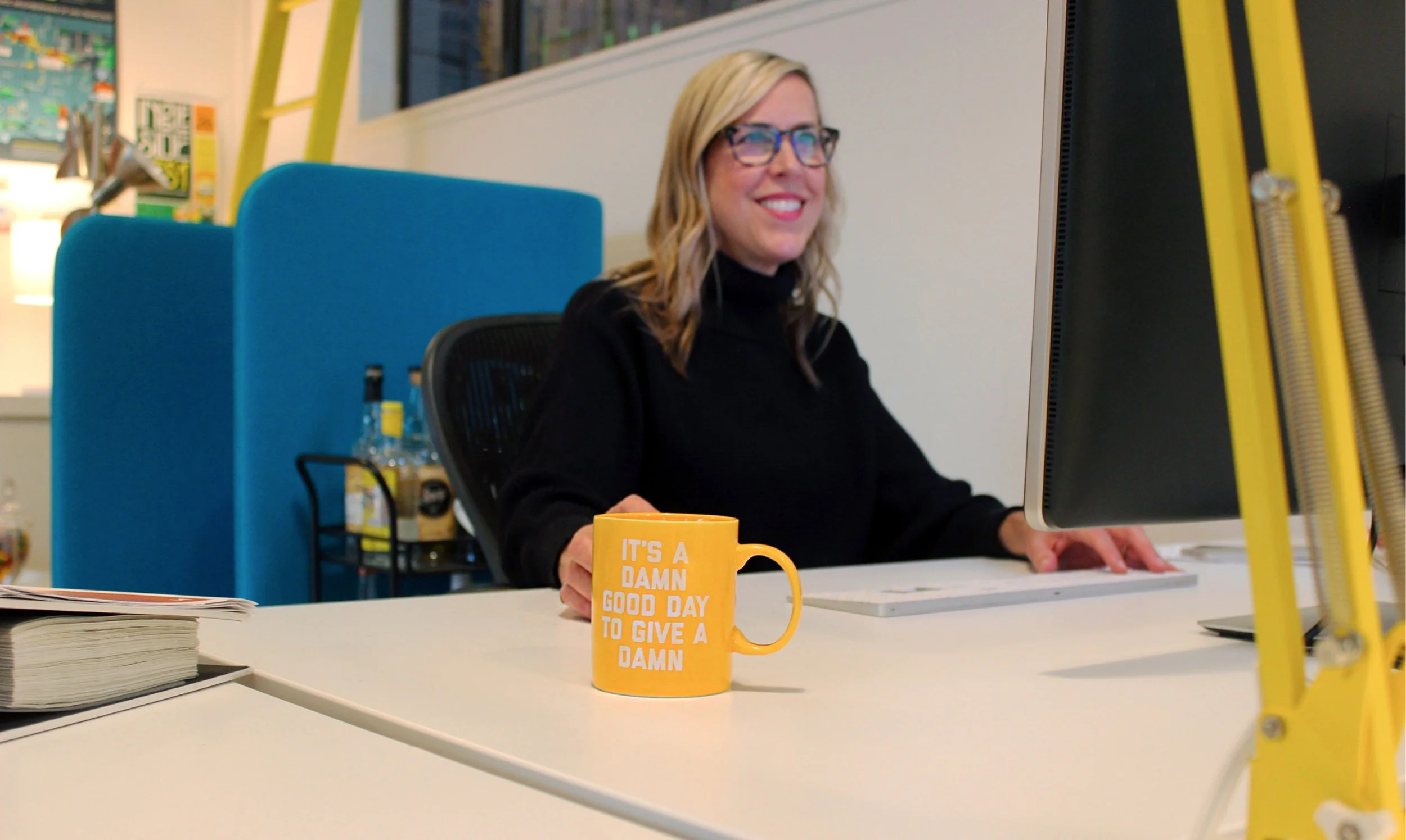 Aimee Kilmer sitting at her desk looking at her computer and smiling with a yellow mug