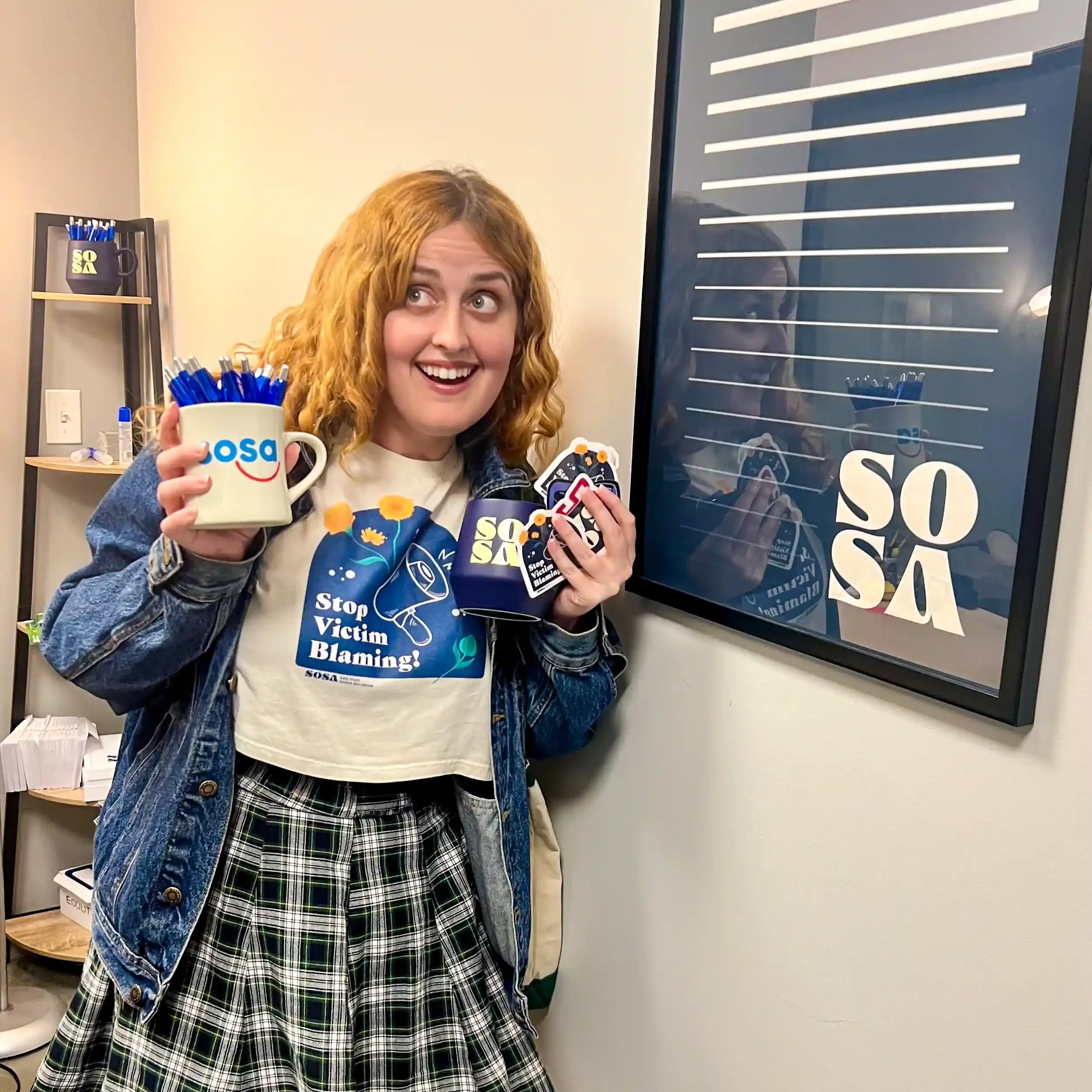 Rachel poses for a photo holding various merchandise (mugs, pens, stickers) next to a poster that reads "SOSA".