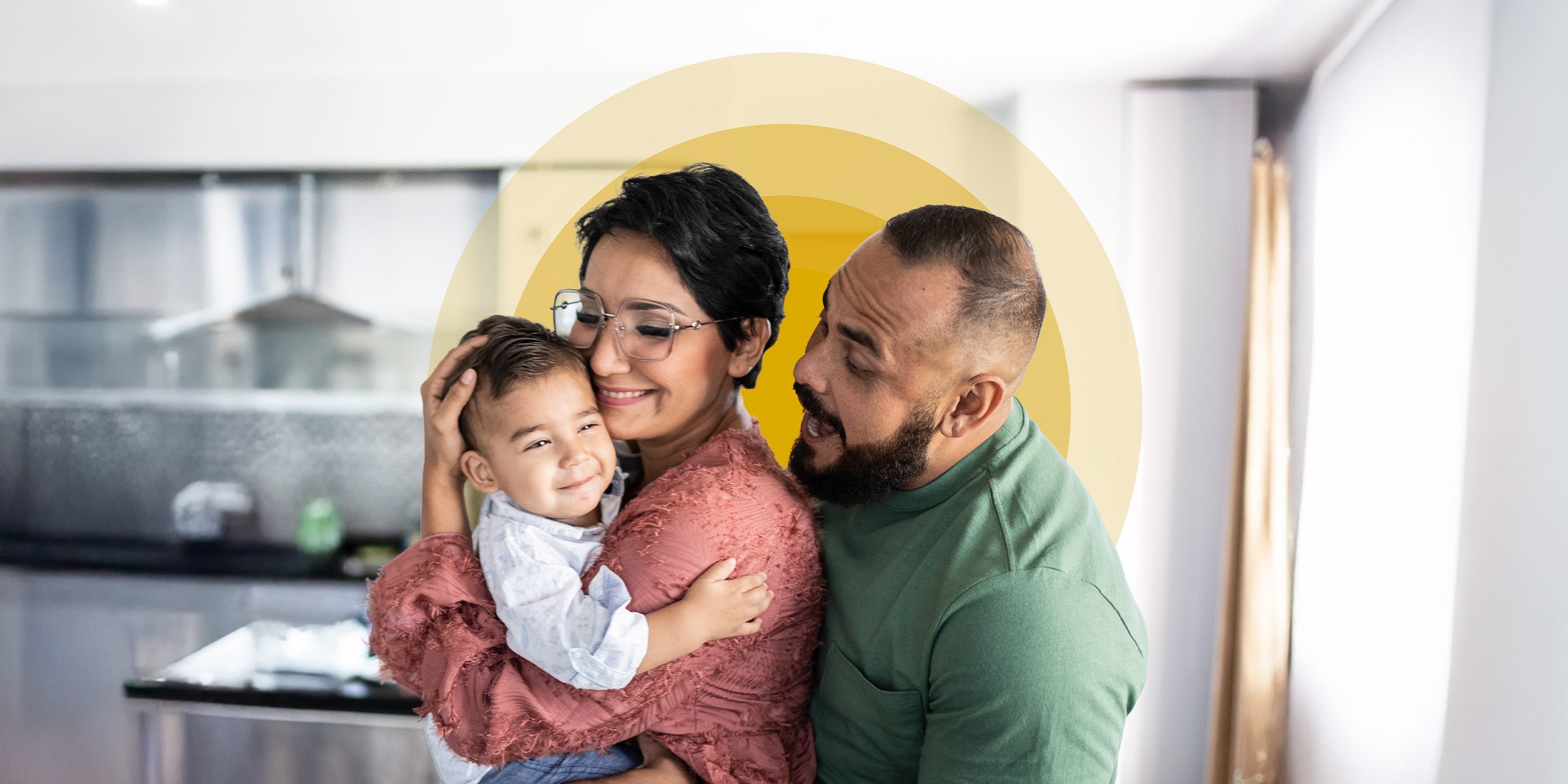 Mother, father, and young son hugging in the kitchen with sun graphic treatment behind them.