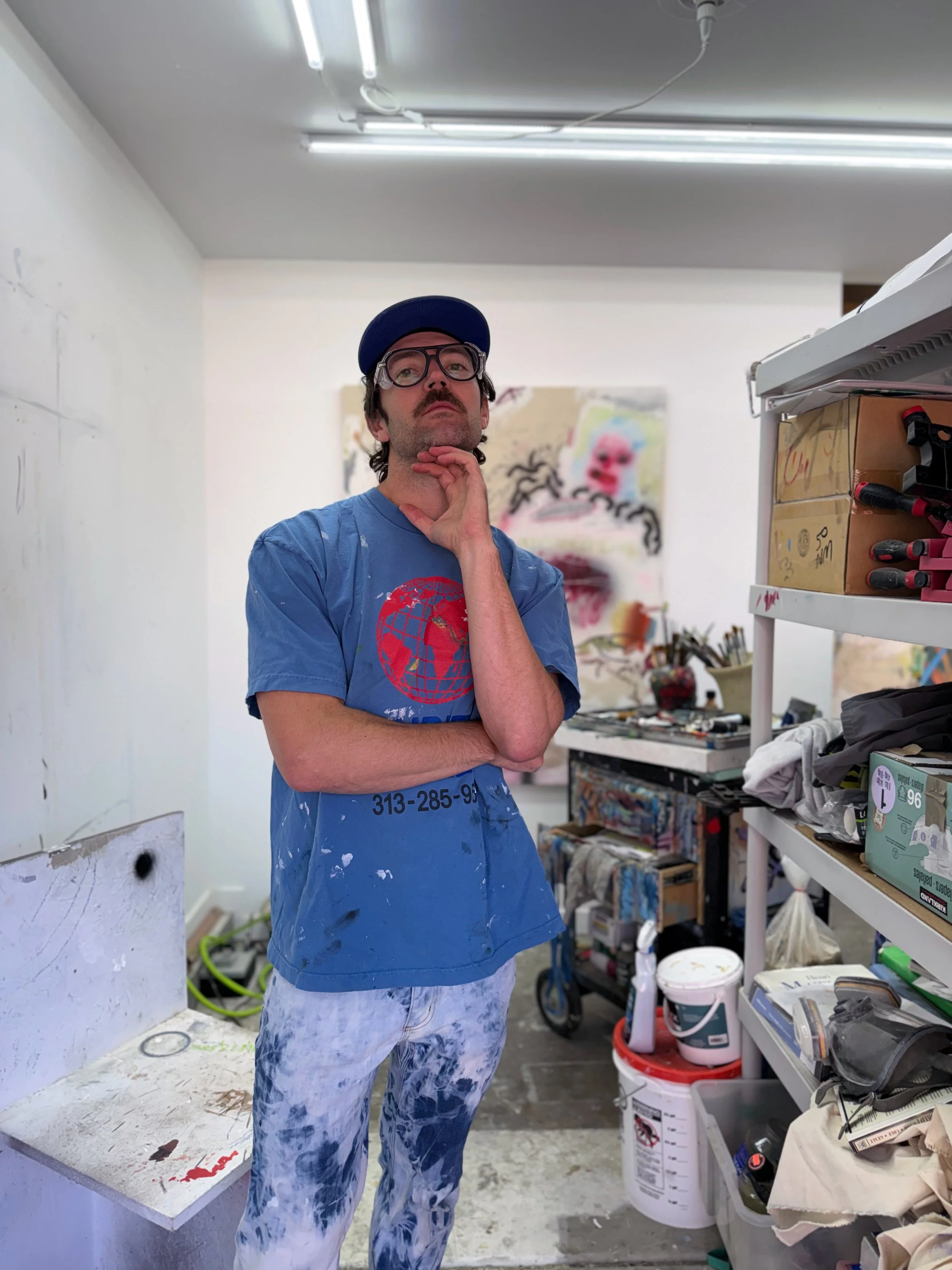 A man in painter's clothes and glasses stands in an art studio, thoughtfully looking upwards with his hand on his chin, surrounded by art supplies and artwork.