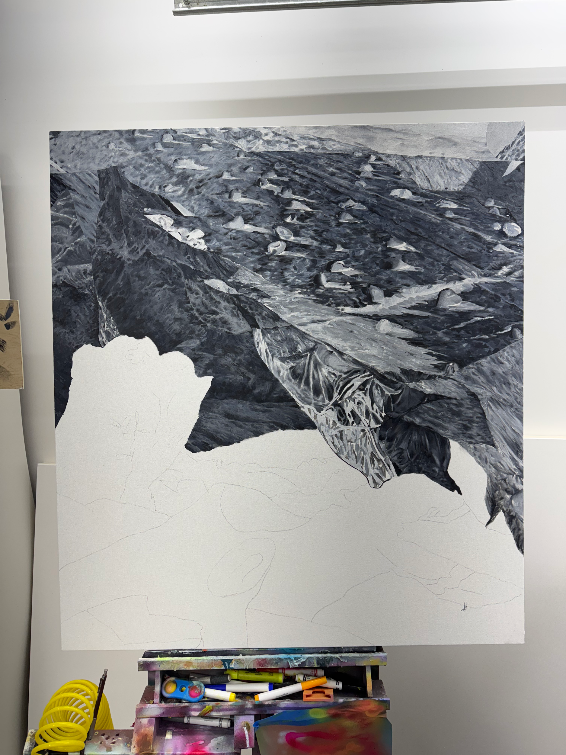 A large painting of a mountain landscape in black and white in progress, with part of the canvas still unpainted, mounted on an easel, which has paint markers and brushes on a colorful palette below.