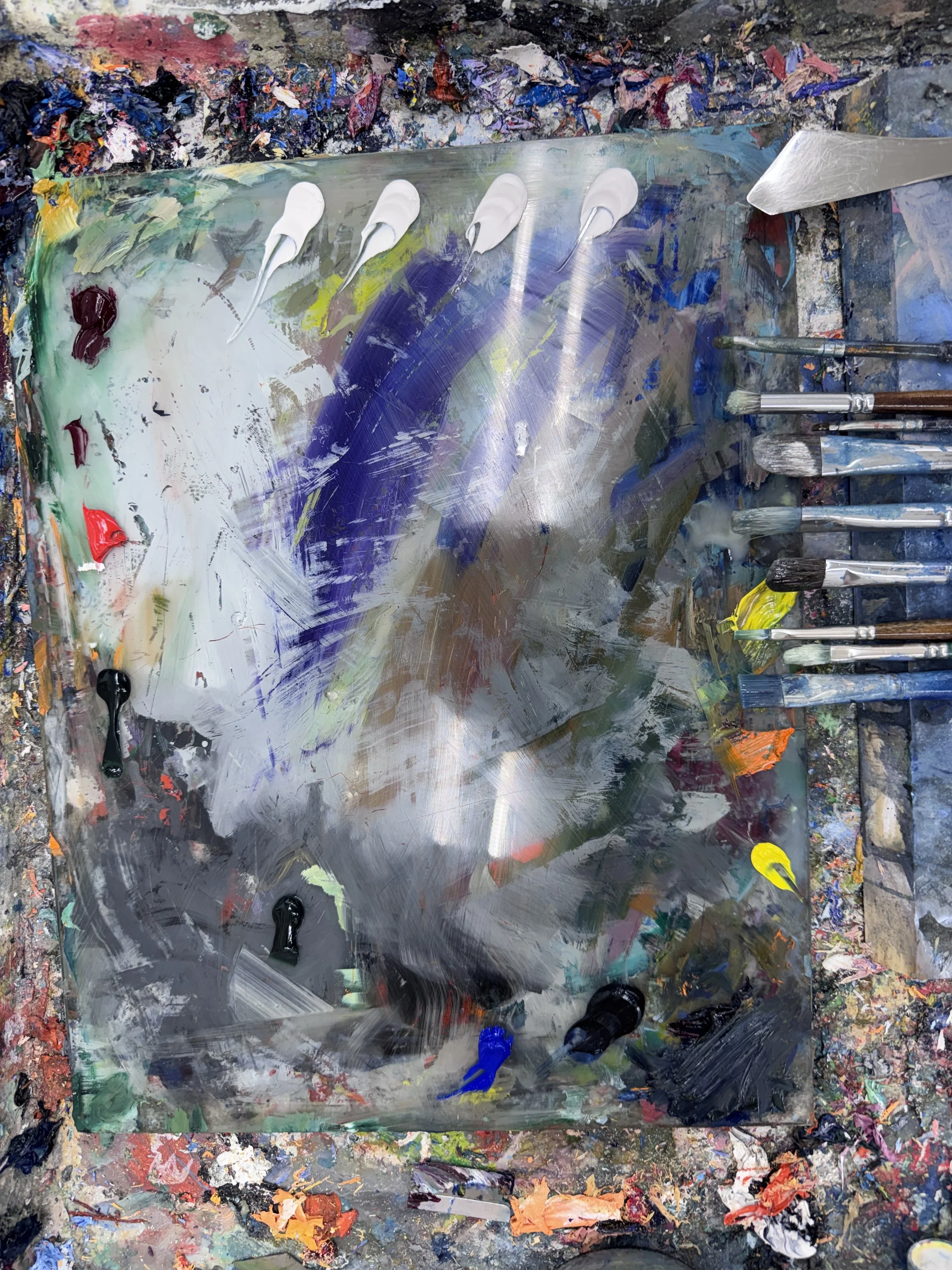 An artist's palette with several blobs of white paint, surrounded by various other colors of paint and brushes, on a paint-splattered surface.