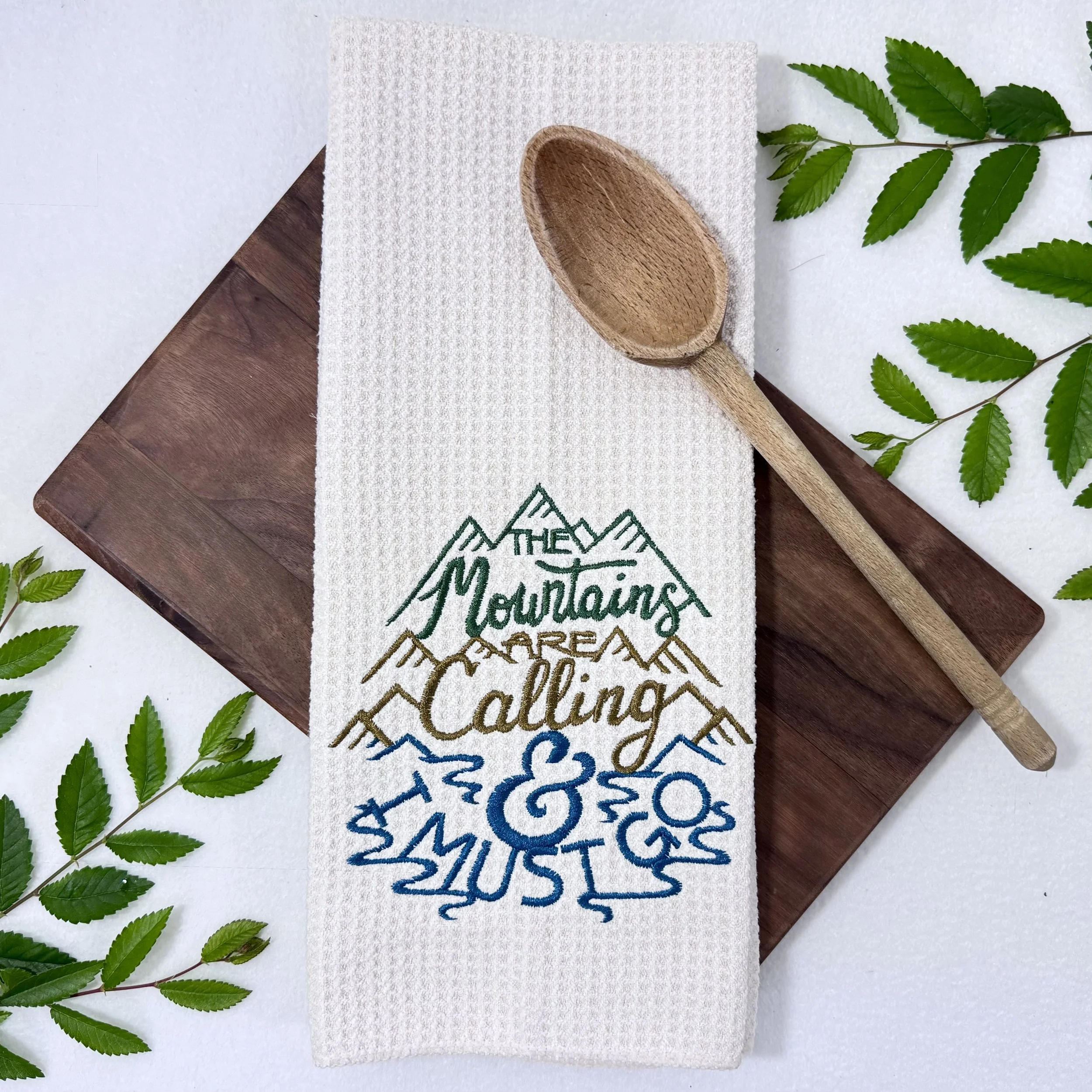 The Mountains Are Calling & I Must Go embroidered microfiber kitchen and hand towel flat lay with greenery and kitchen tools