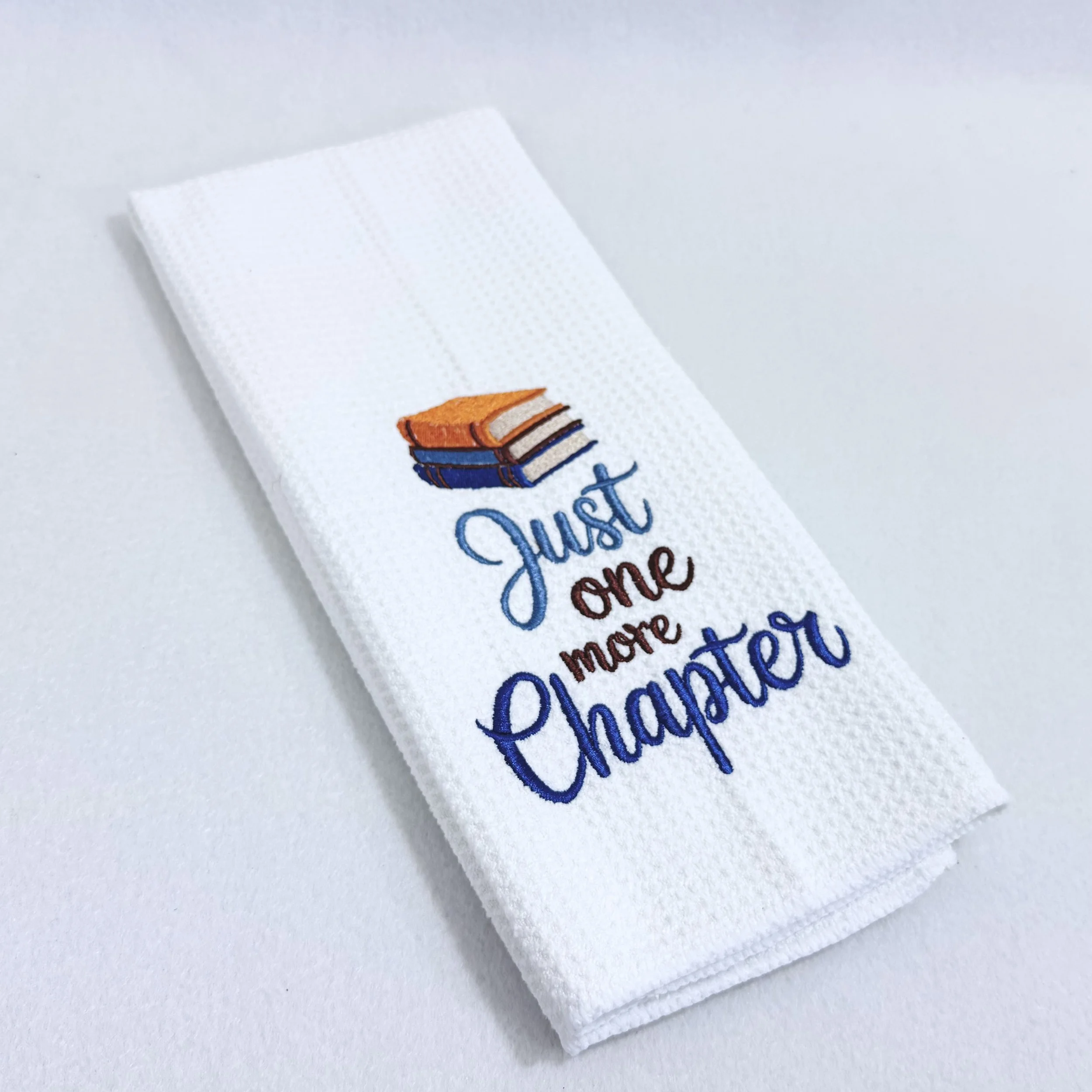 The Just One More Chapter embroidered microfiber kitchen and hand towel folded on white background, showing books design and texture