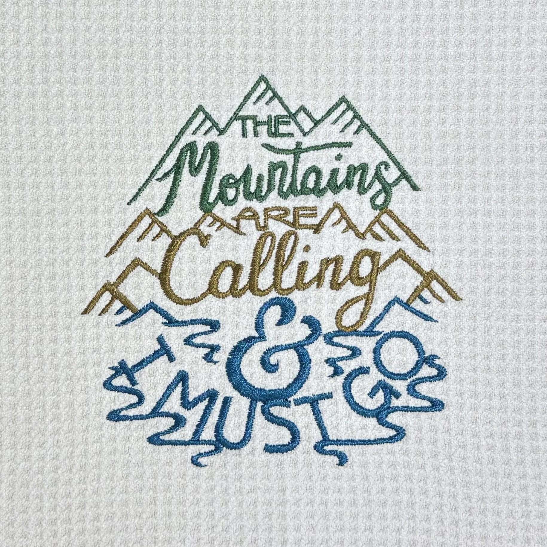 Close-up of The Mountains Are Calling & I Must Go embroidery on microfiber kitchen and hand towel, showing detailed stitching, mountain design, and towel texture