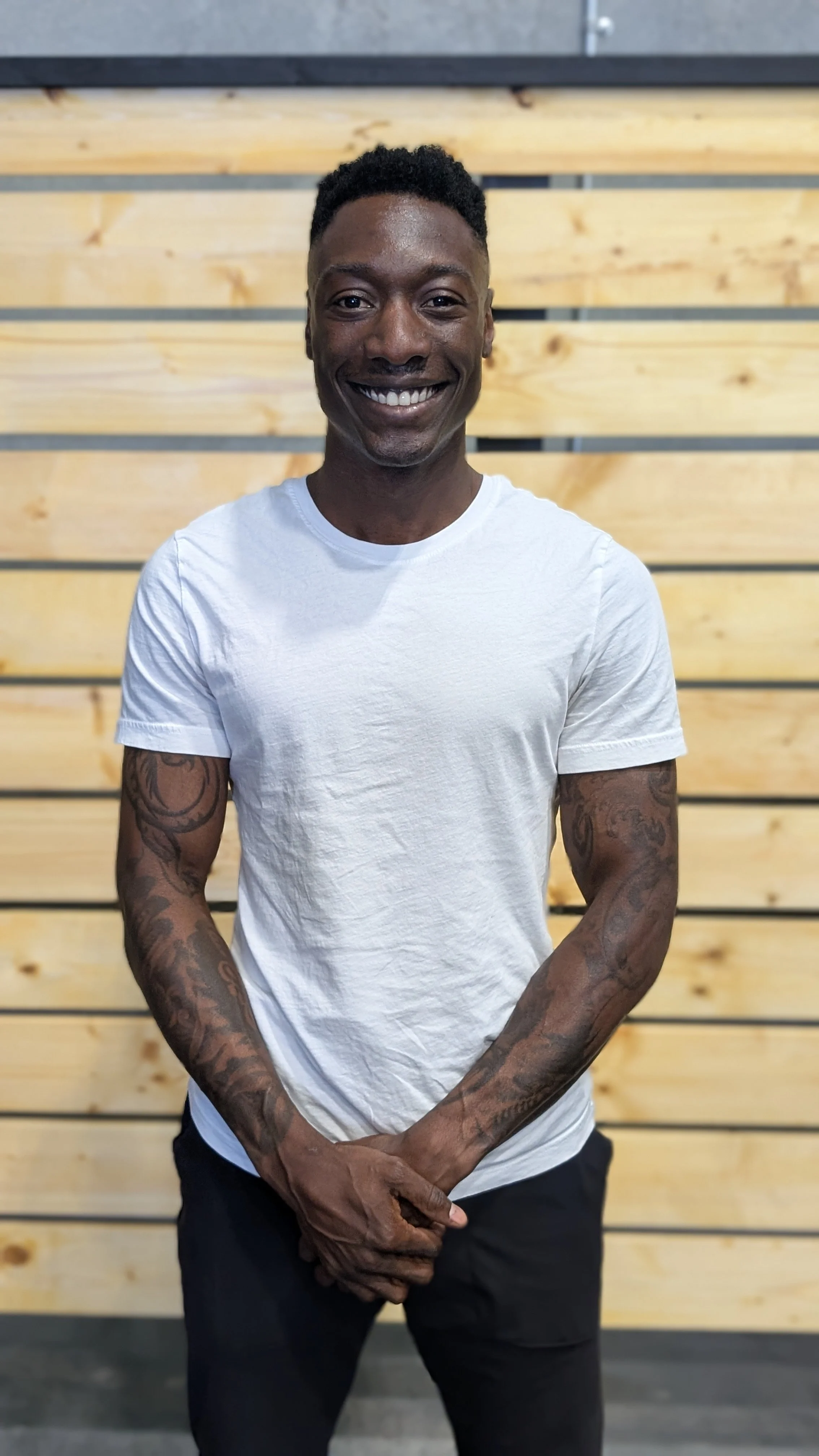 Smiling man with tattoos on arms, wearing a white t-shirt, standing in front of a wooden wall.
