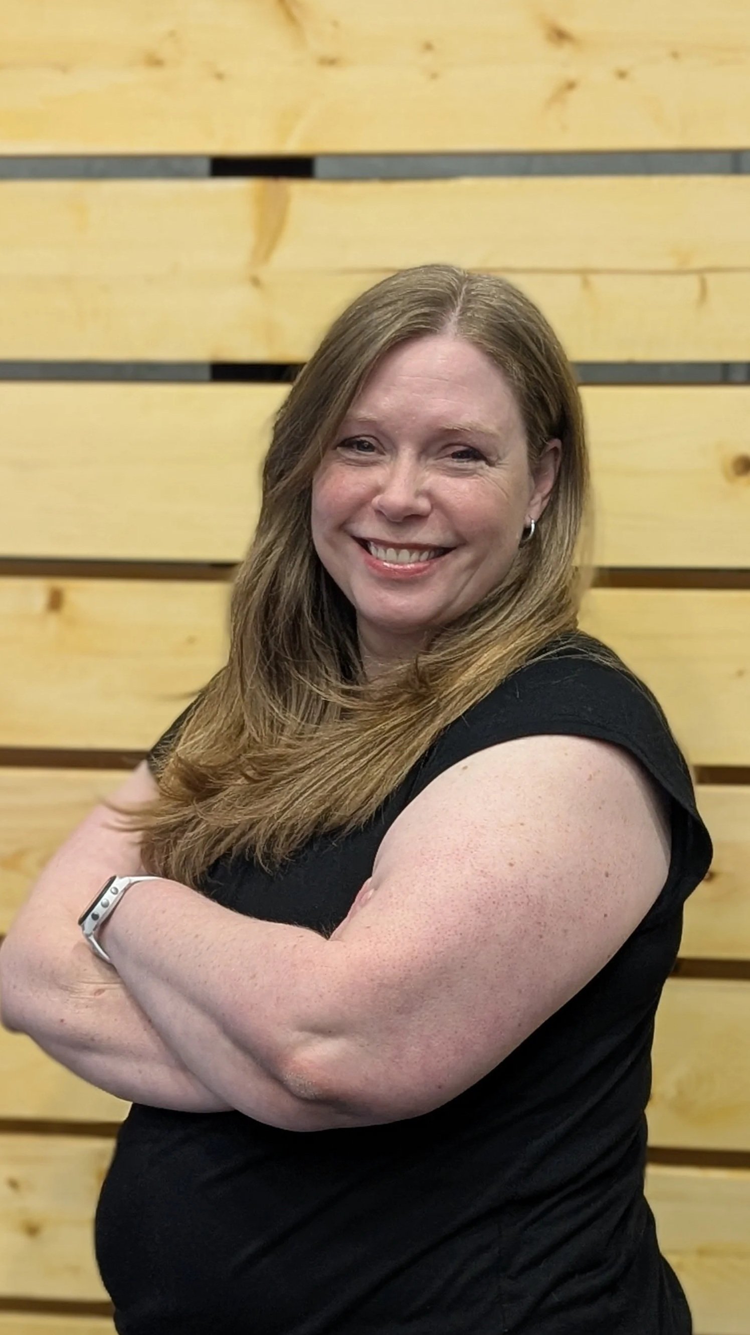 A woman with long, wavy, light brown hair smiling with arms crossed in front of a wooden panel background.