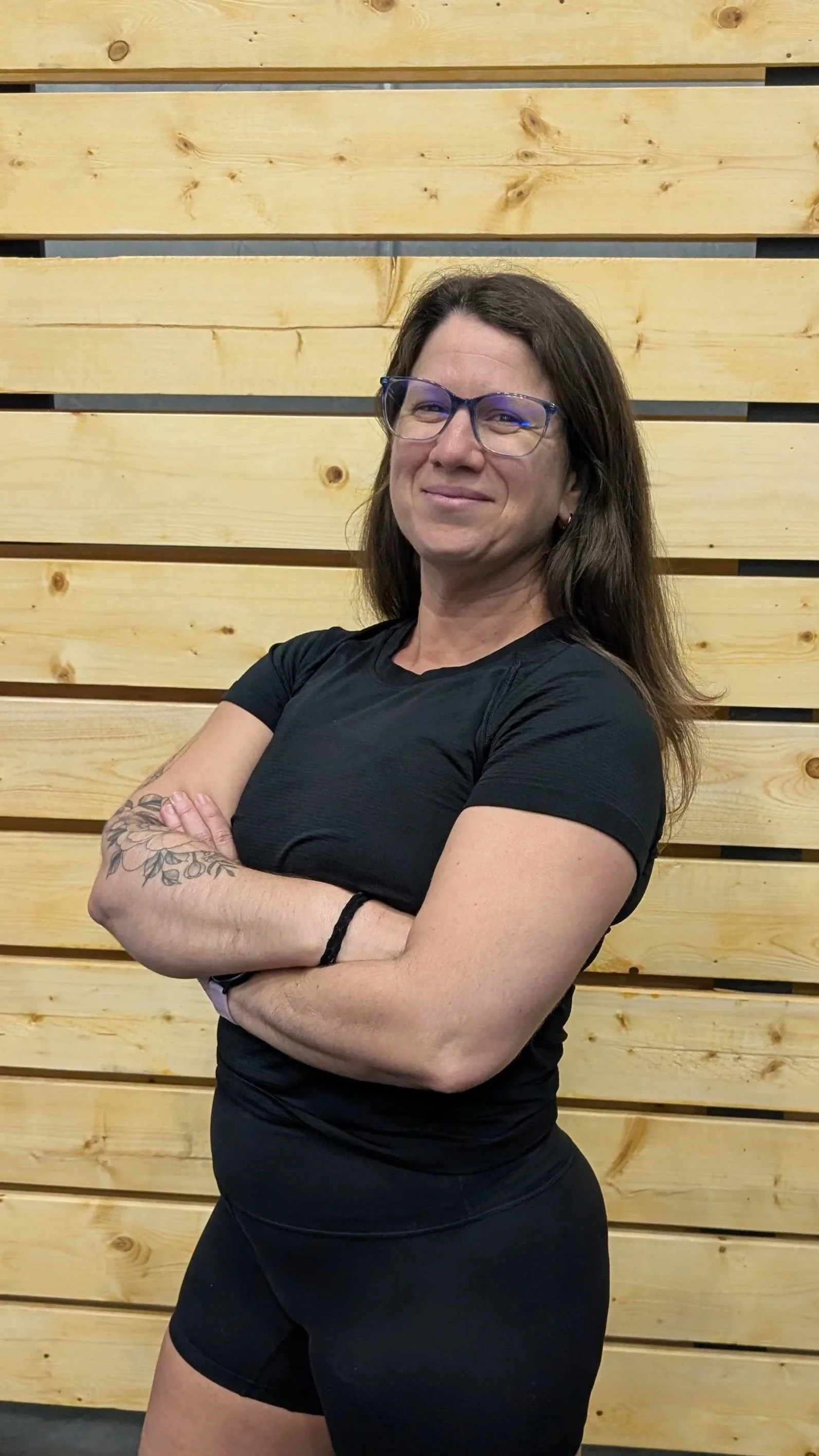 A woman with glasses, long brown hair, and tattoos on her right arm, wearing a black athletic shirt and shorts, standing with her arms crossed in front of a wooden wall.