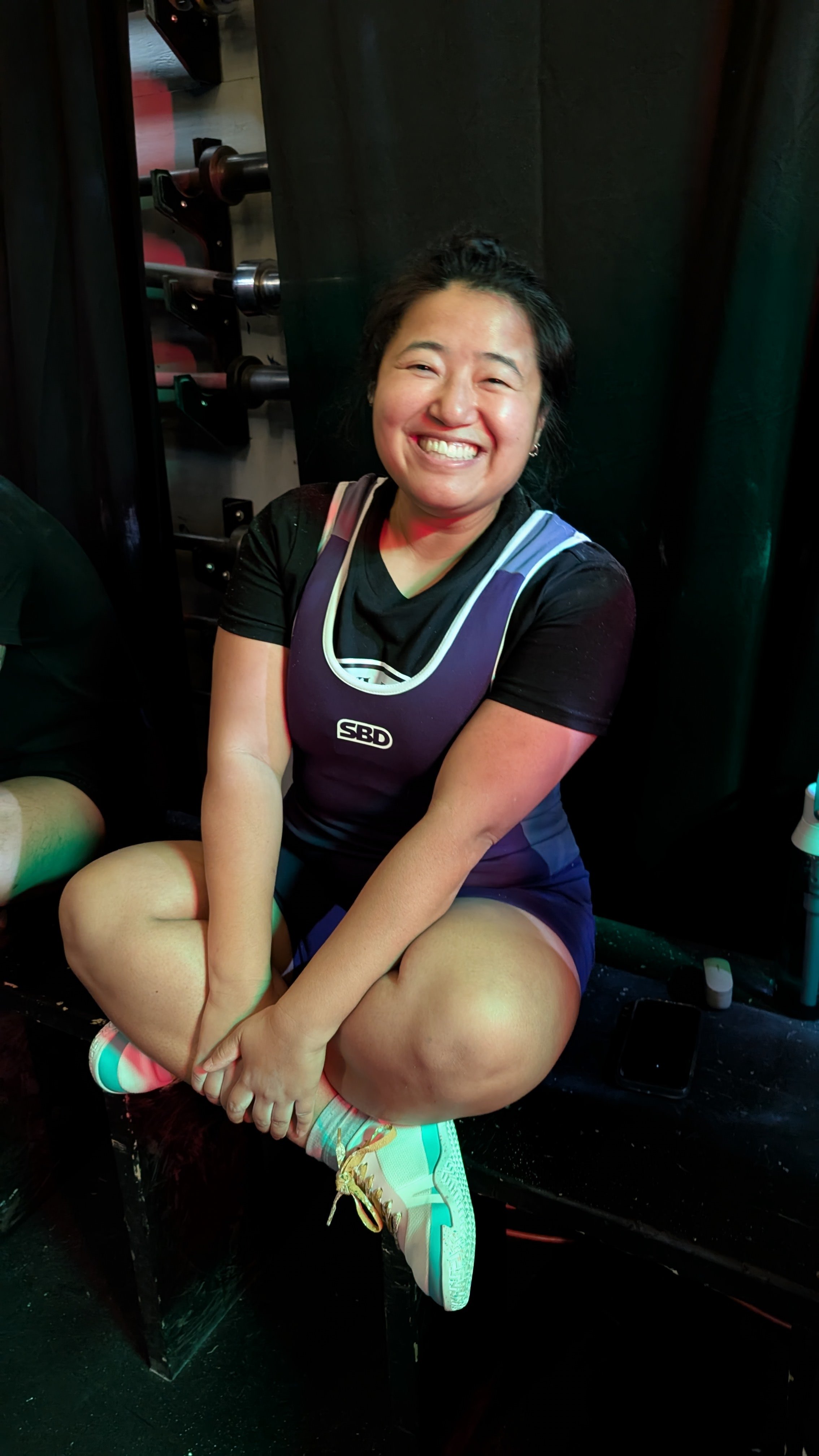 Smiling woman sitting cross-legged on a bench in a gym or weightlifting area, wearing a purple powerlifting singlet, black t-shirt, colorful sneakers, and surrounded by fitness equipment.