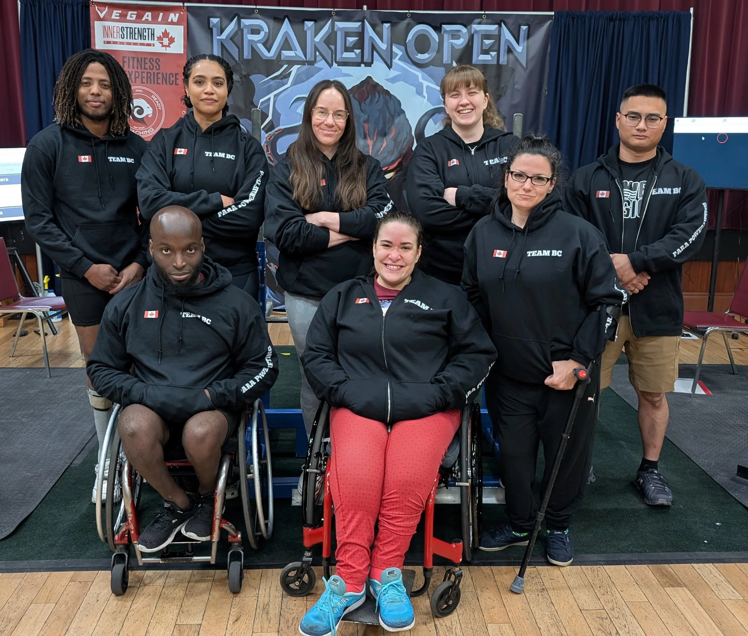 Group of eight people, some in wheelchair, wearing black hoodies with Canadian flags and 'TEAM BC' printed, standing and sitting on a stage with a backdrop that says 'KRAKEN OPEN'.