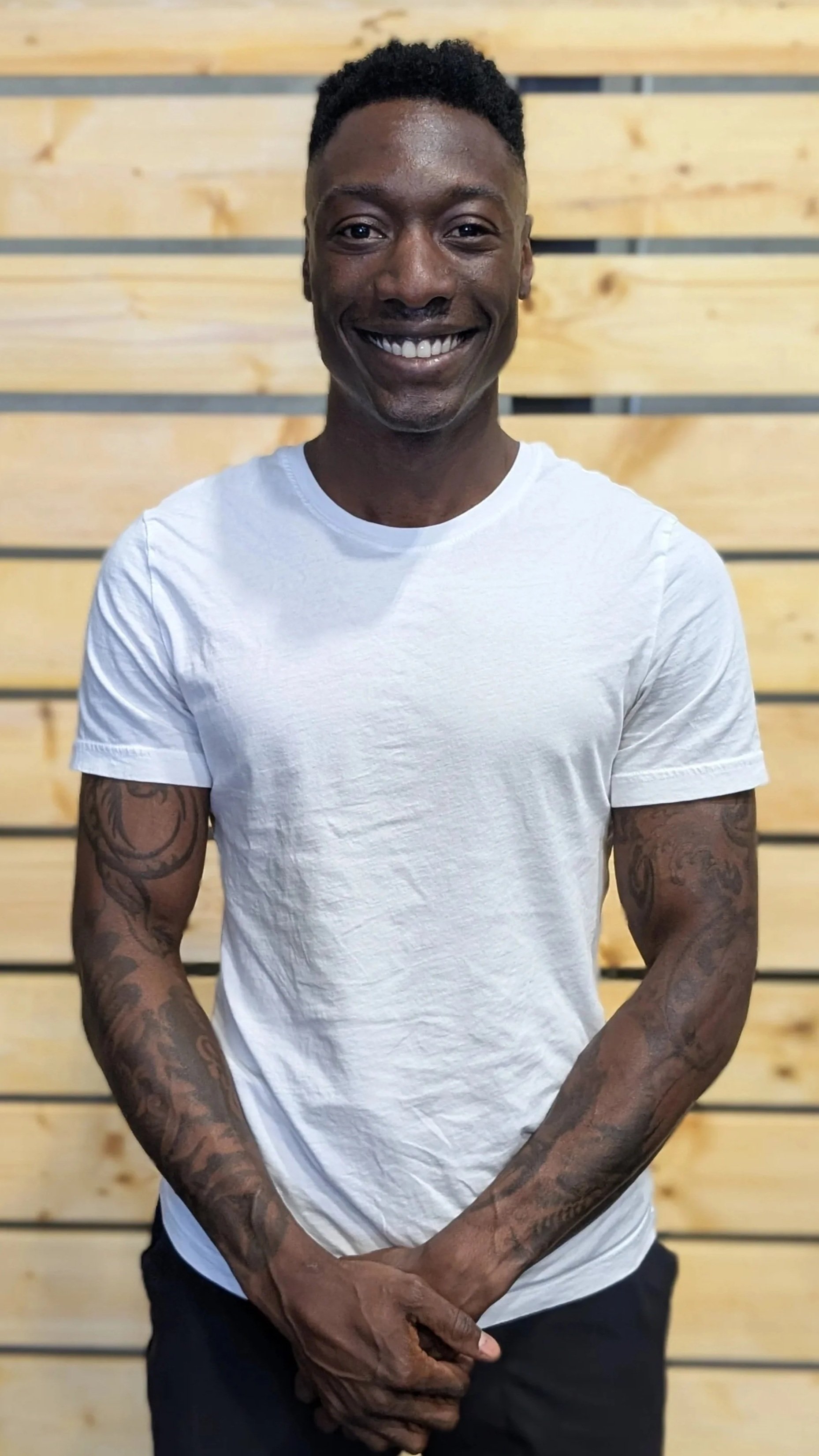 Smiling man with short curly hair and tattoos on arms, wearing a white t-shirt, standing in front of a wooden slatted wall.