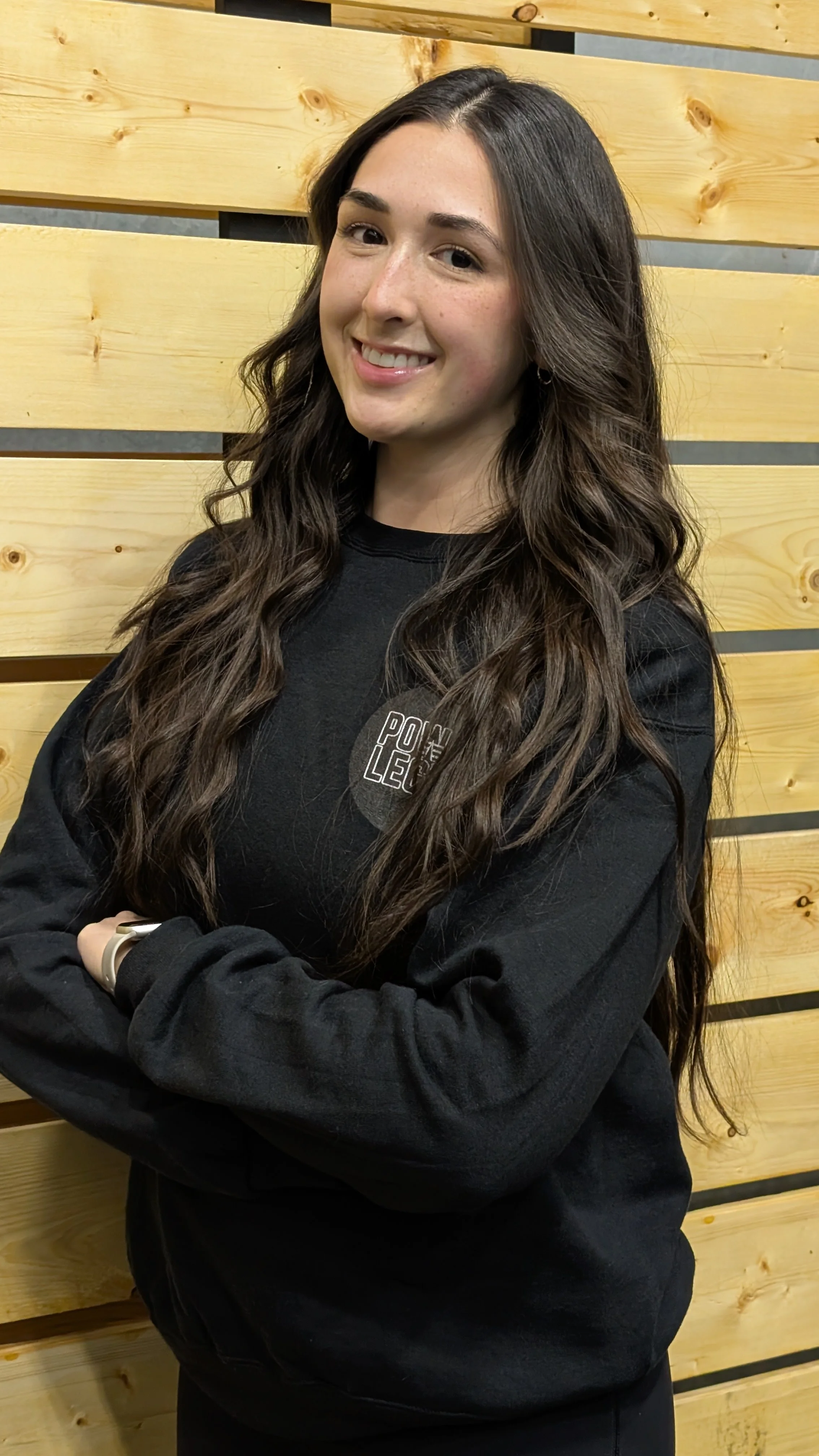 A young woman with long, wavy brown hair smiling and standing with arms crossed in front of a wooden wall.