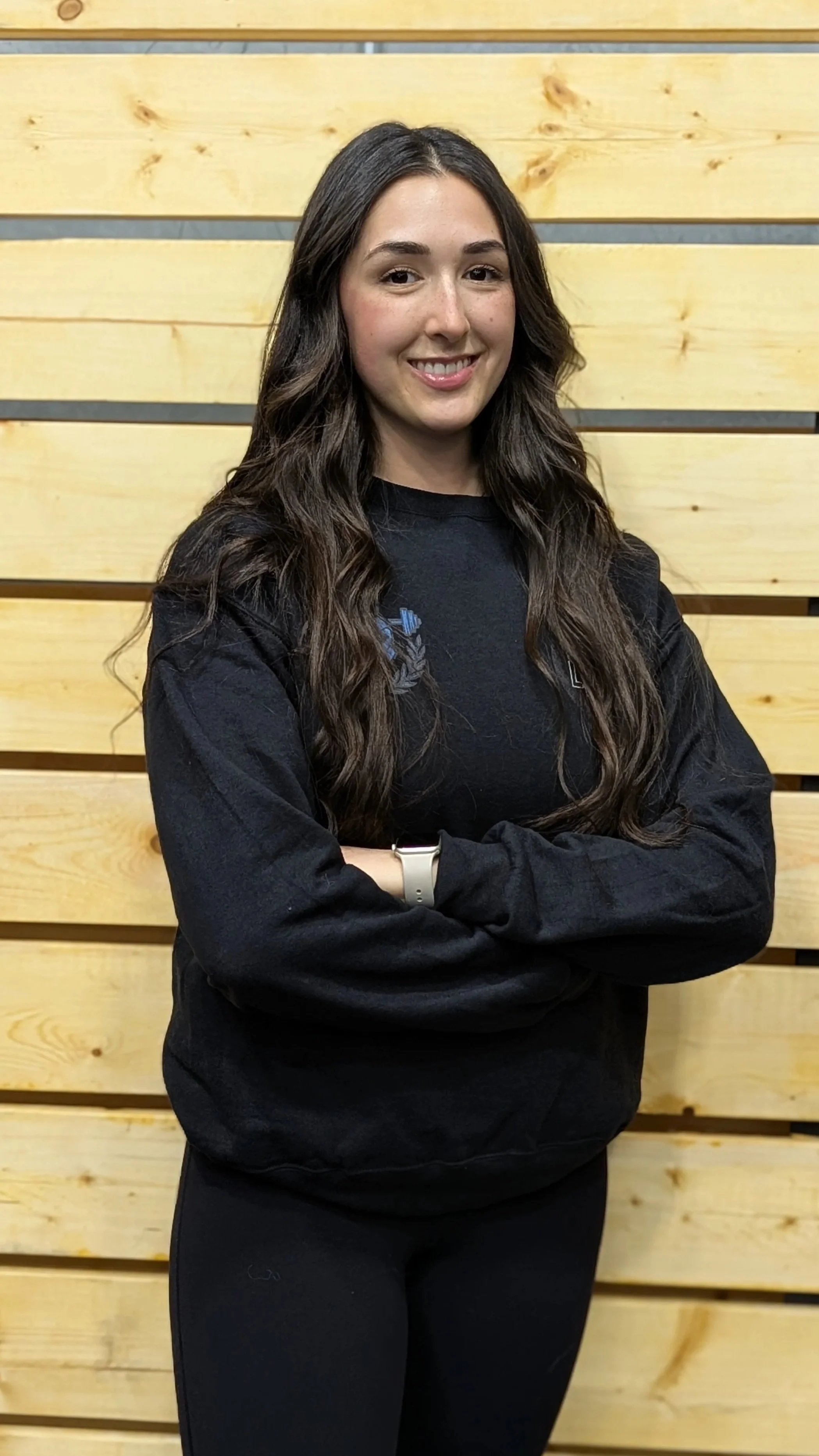 A young woman with long dark hair, smiling and crossing her arms, standing against a wooden wall.