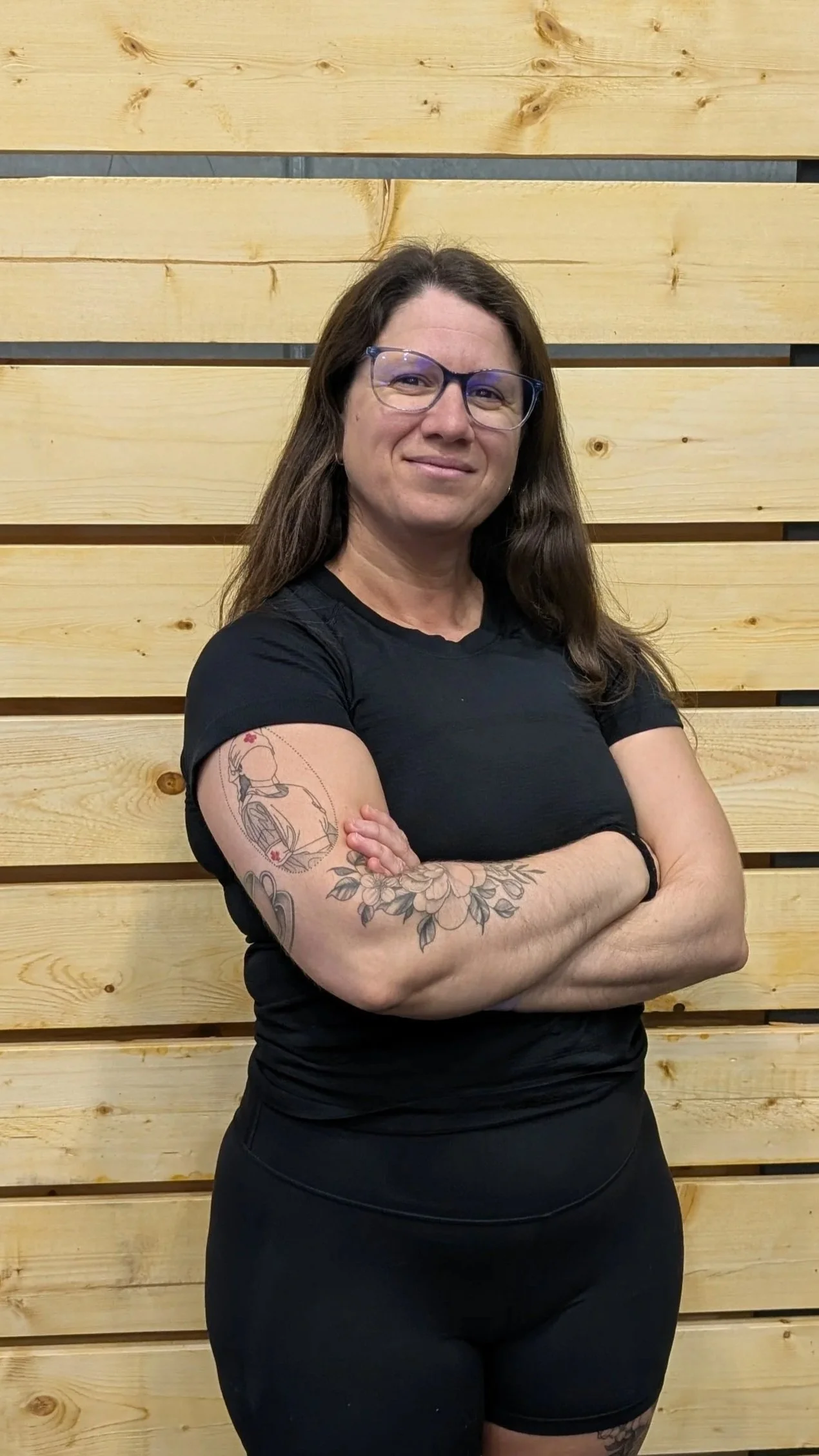 A woman with long dark hair, glasses, and tattooed arms standing with arms crossed in front of a wooden wall.