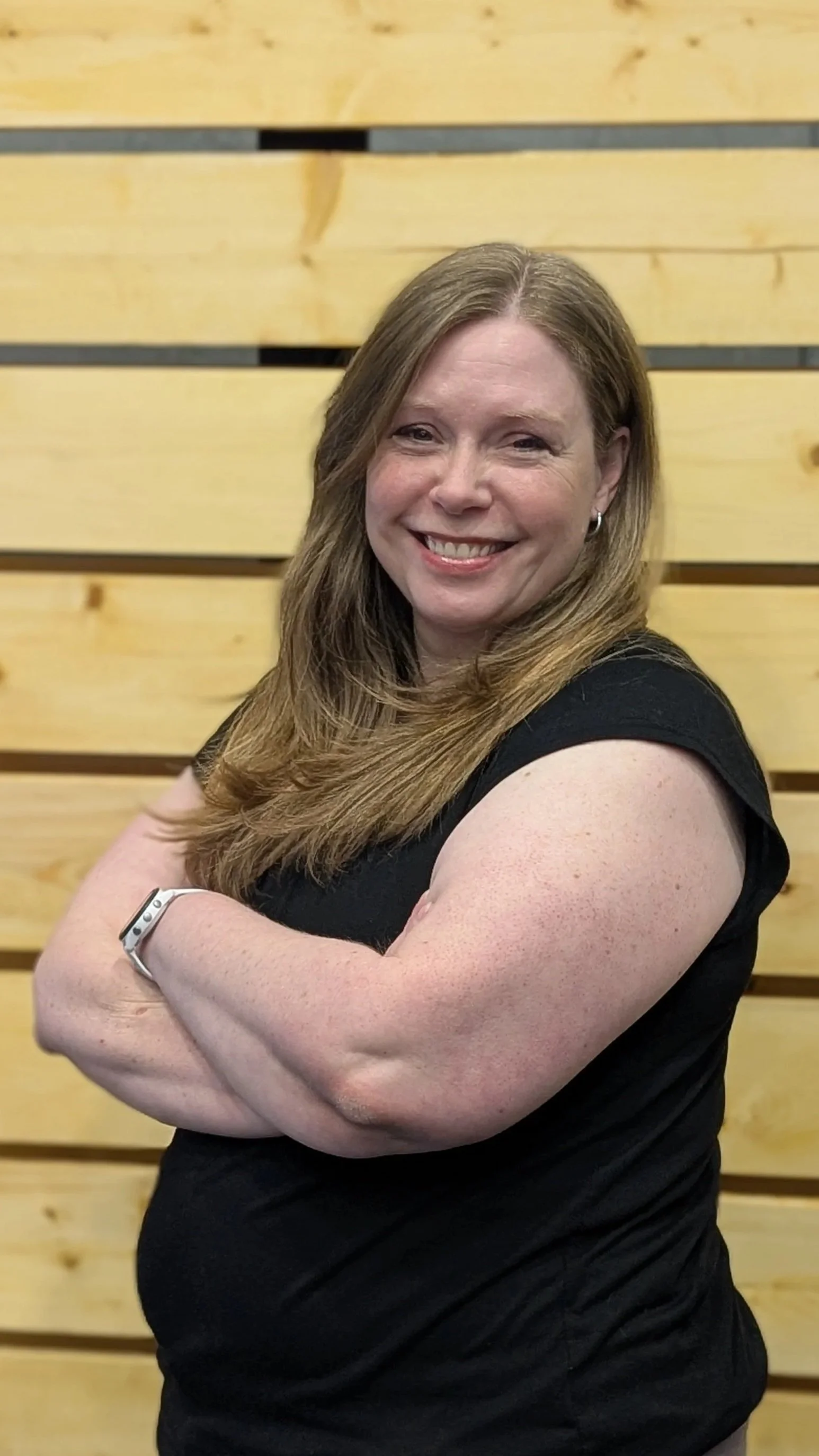 Photo of a smiling woman with long, light brown hair, wearing a black short-sleeved top, standing in front of a wooden wall with horizontal planks.