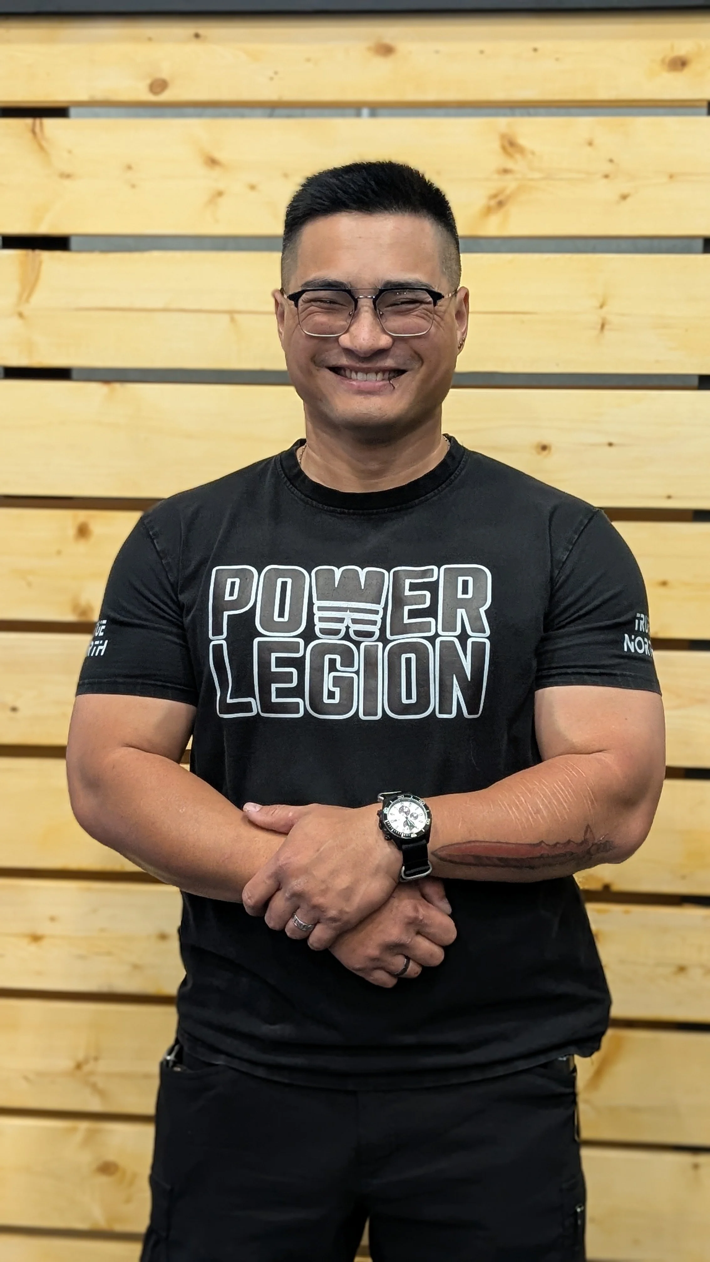 A smiling man with short black hair, glasses, and a muscular build, wearing a black Power of the Legion T-shirt, clasping his hands in front of him, standing in front of a light wooden wall.