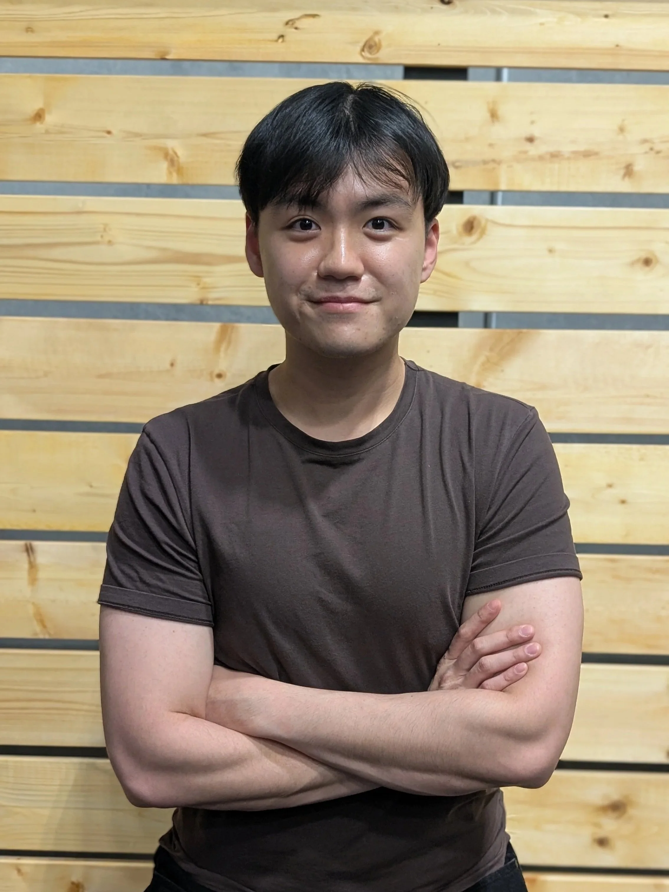 A young man with black hair and light skin standing against a wooden wall, wearing a dark T-shirt, arms crossed, smiling subtly.
