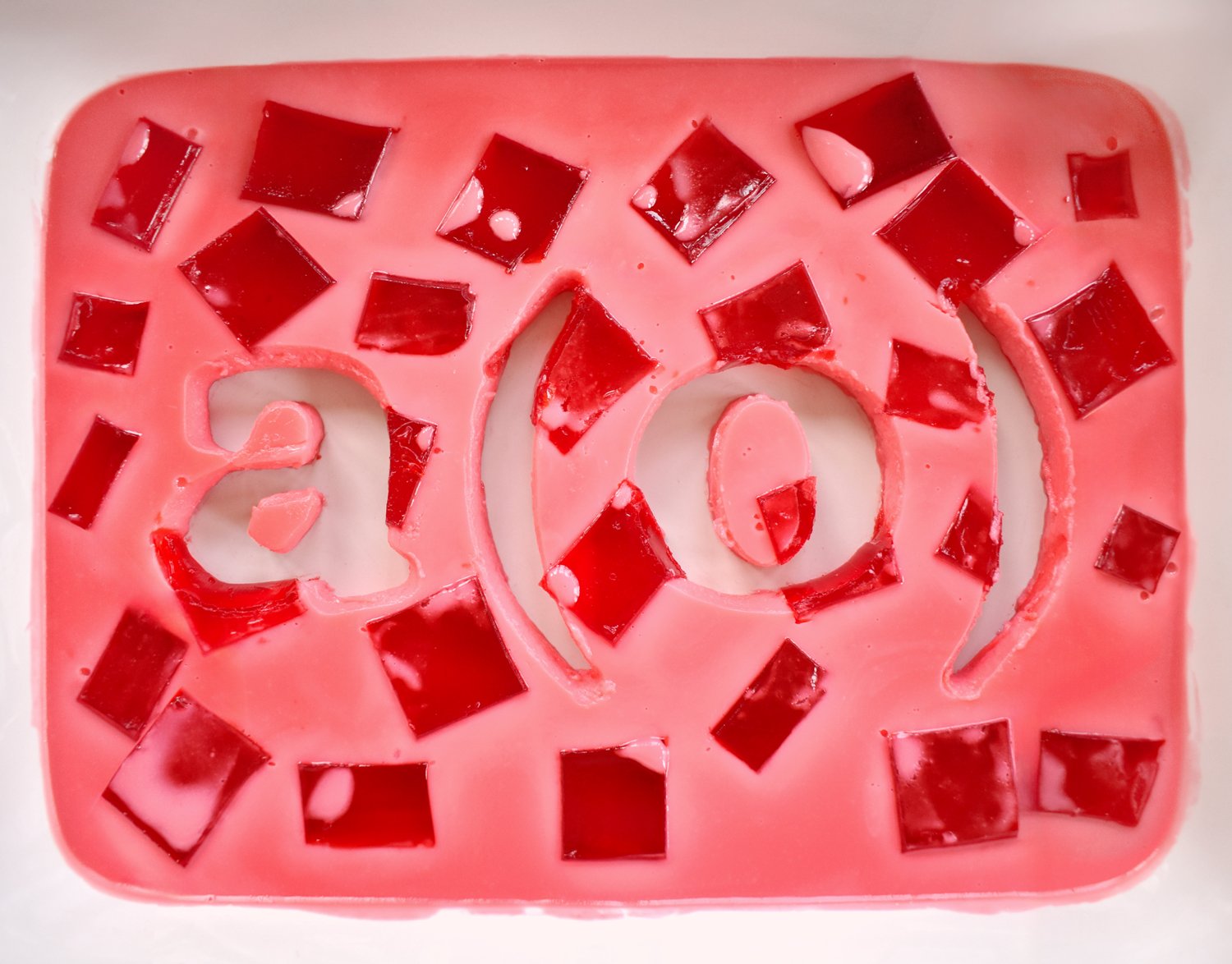 A cutout of the A(O) logo, surrounded by a rectangular field of milky pink gelatin and scattered red gelatin pieces, revealing a white ceramic dish below.
