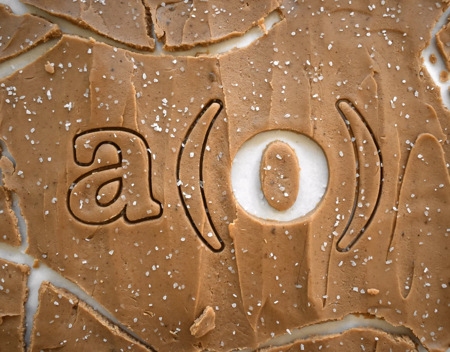 A sheet of sea-salted dulce de leche bark inscribed with the "a(o)" logo. Cracks and missing pieces reveal parchment baking paper underneath.