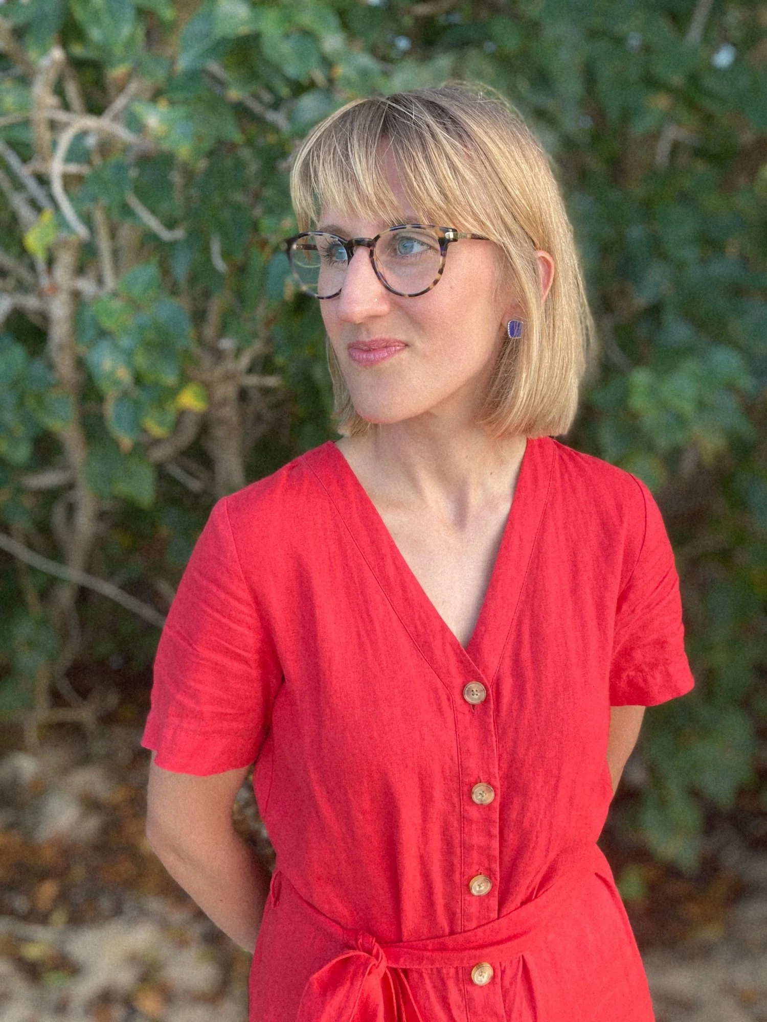 Nicole, A(O)'s Co-founder, standing in front of trees with her hands behind her back, smiling gently as she gazes to the left. She is wearing glasses, a red romper and blue stud earrings.