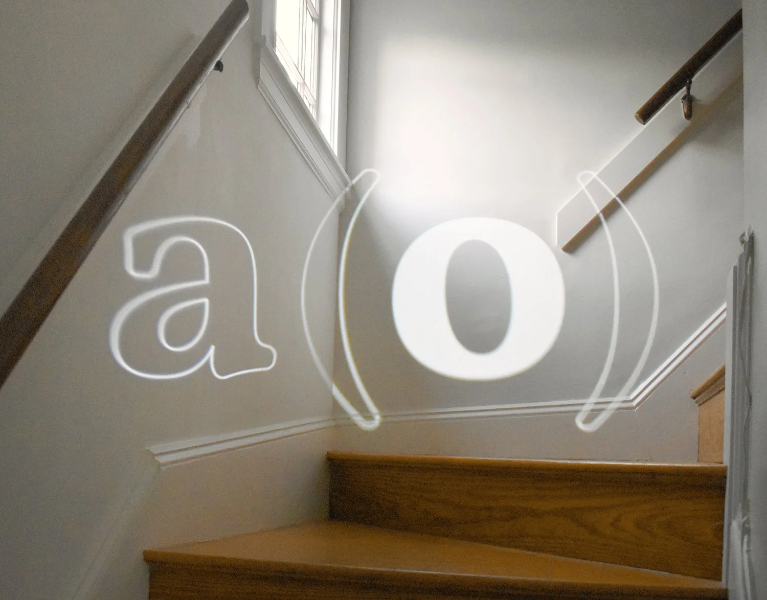 A white light projects the "a(o)" logo onto the walls of a staircase, surrounded by wooden railings and steps.