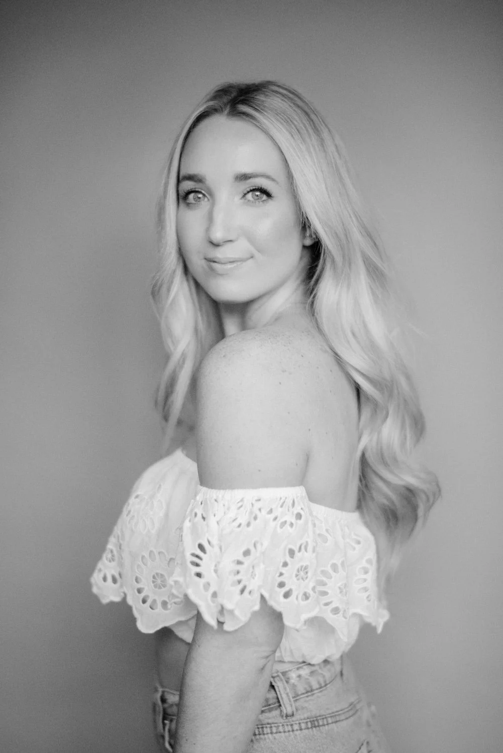 Black and white portrait of a woman with long wavy hair, wearing a pieced off-shoulder lace top and jeans, smiling gently at the camera.