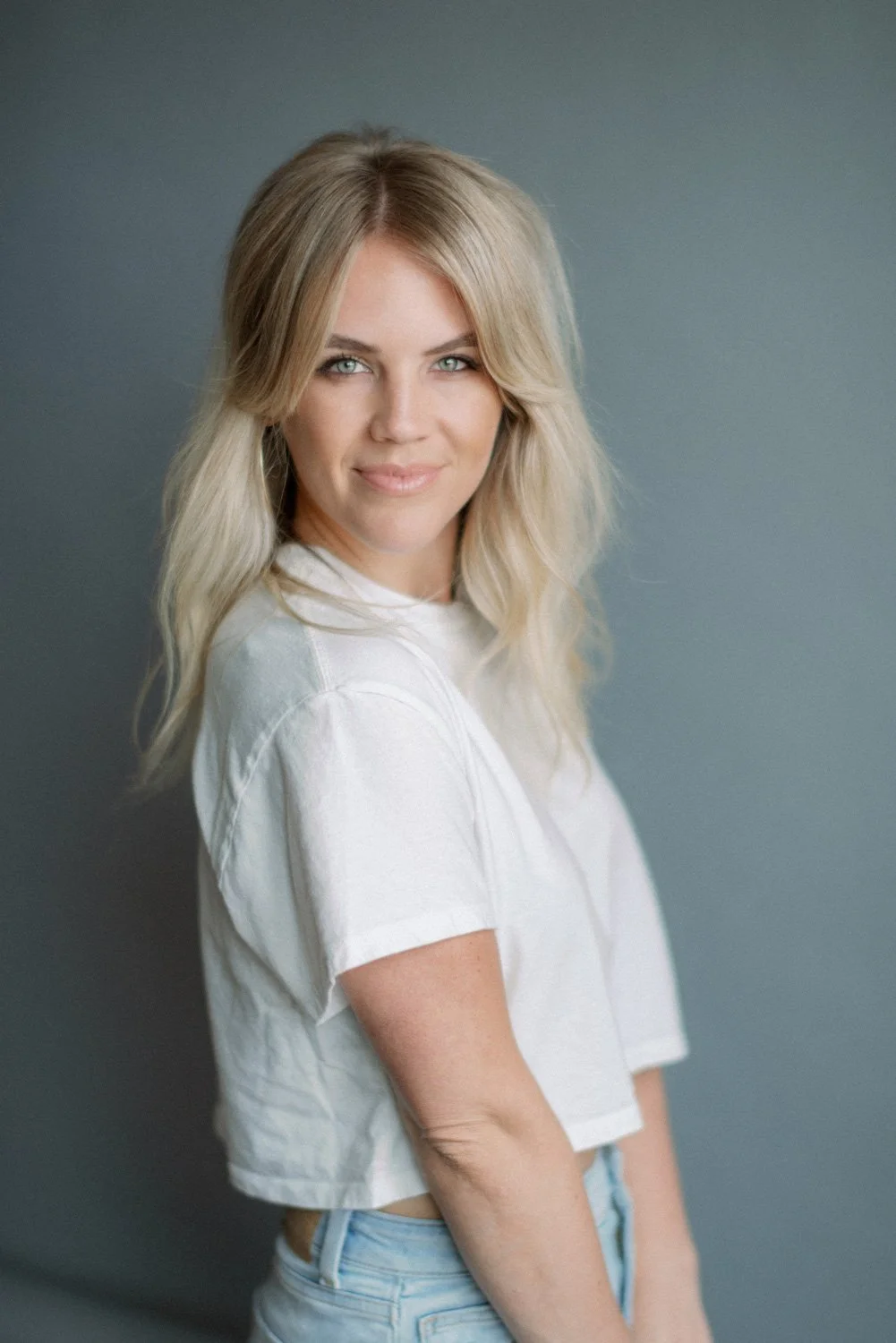 Portrait of a smiling blonde woman with wavy hair, wearing a white t-shirt, standing against a gray background.