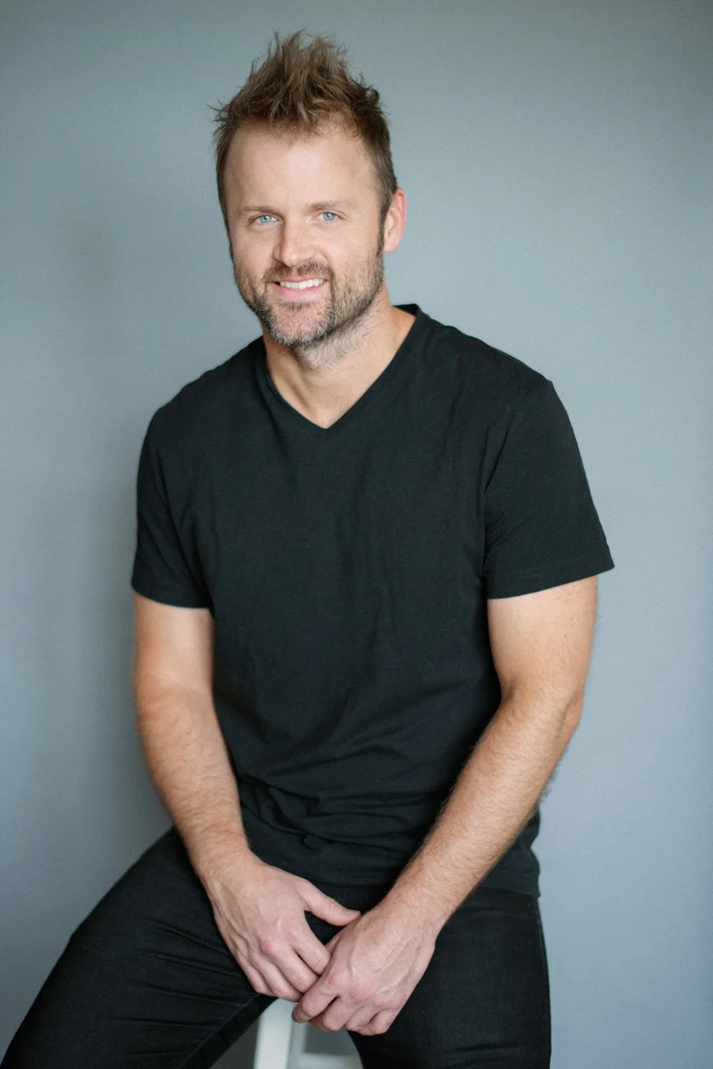 A man with short brown hair and a beard, wearing a black t-shirt, sitting against a gray wall, smiling at the camera.
