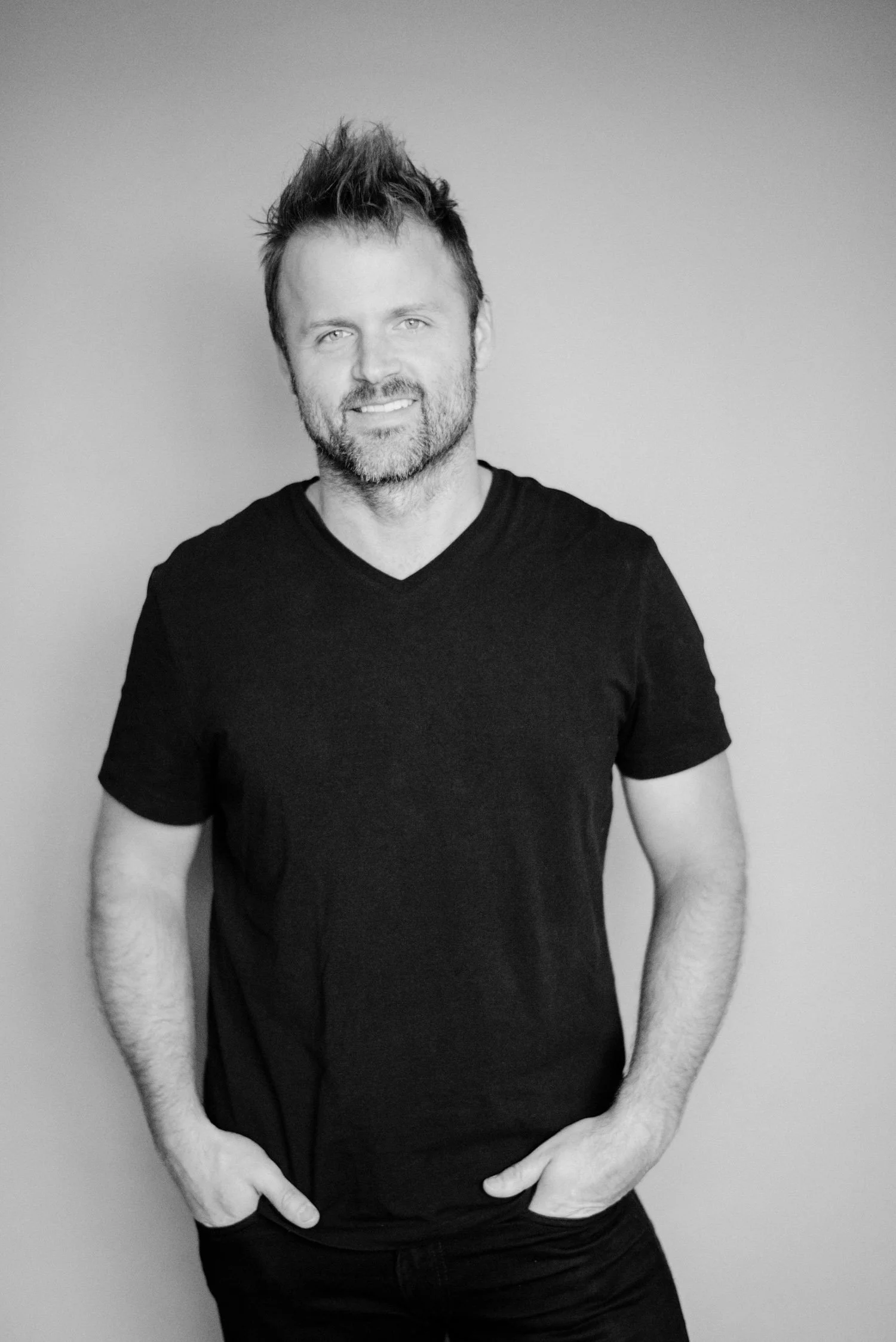 Black and white portrait of a smiling man with light eyes, groomed beard, and short tousled hair, wearing a black V-neck T-shirt, standing against a plain background.
