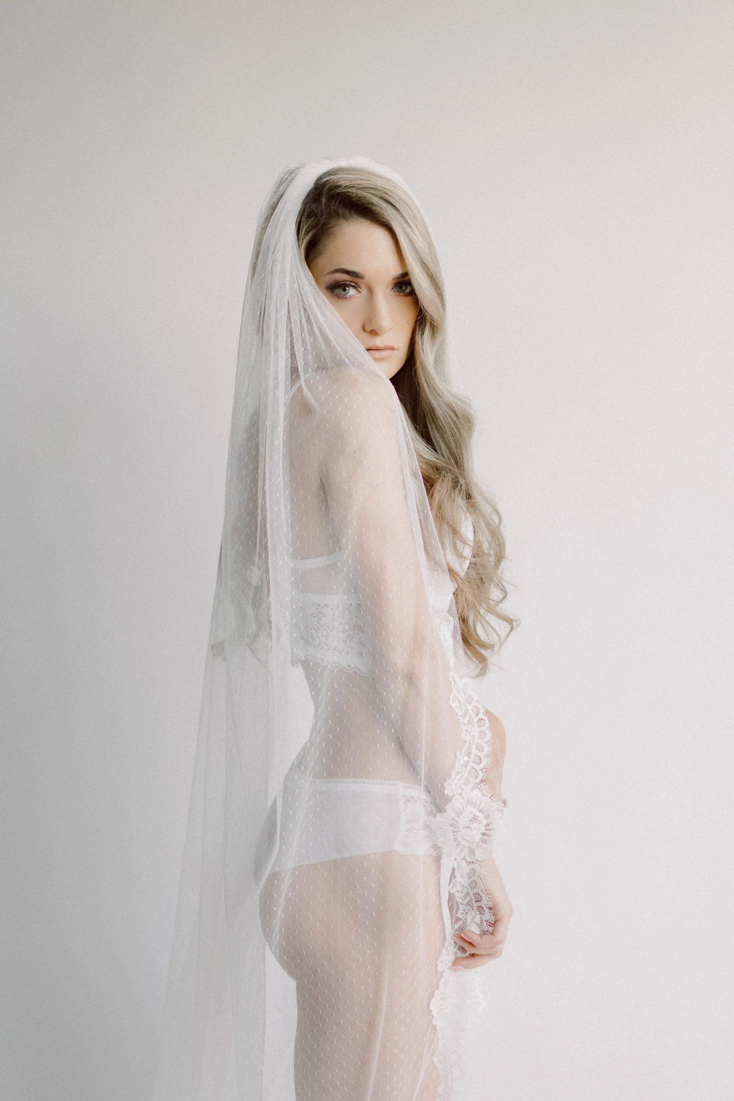 A woman with long wavy hair wearing white lace lingerie and a sheer veil, posing against a plain light background.